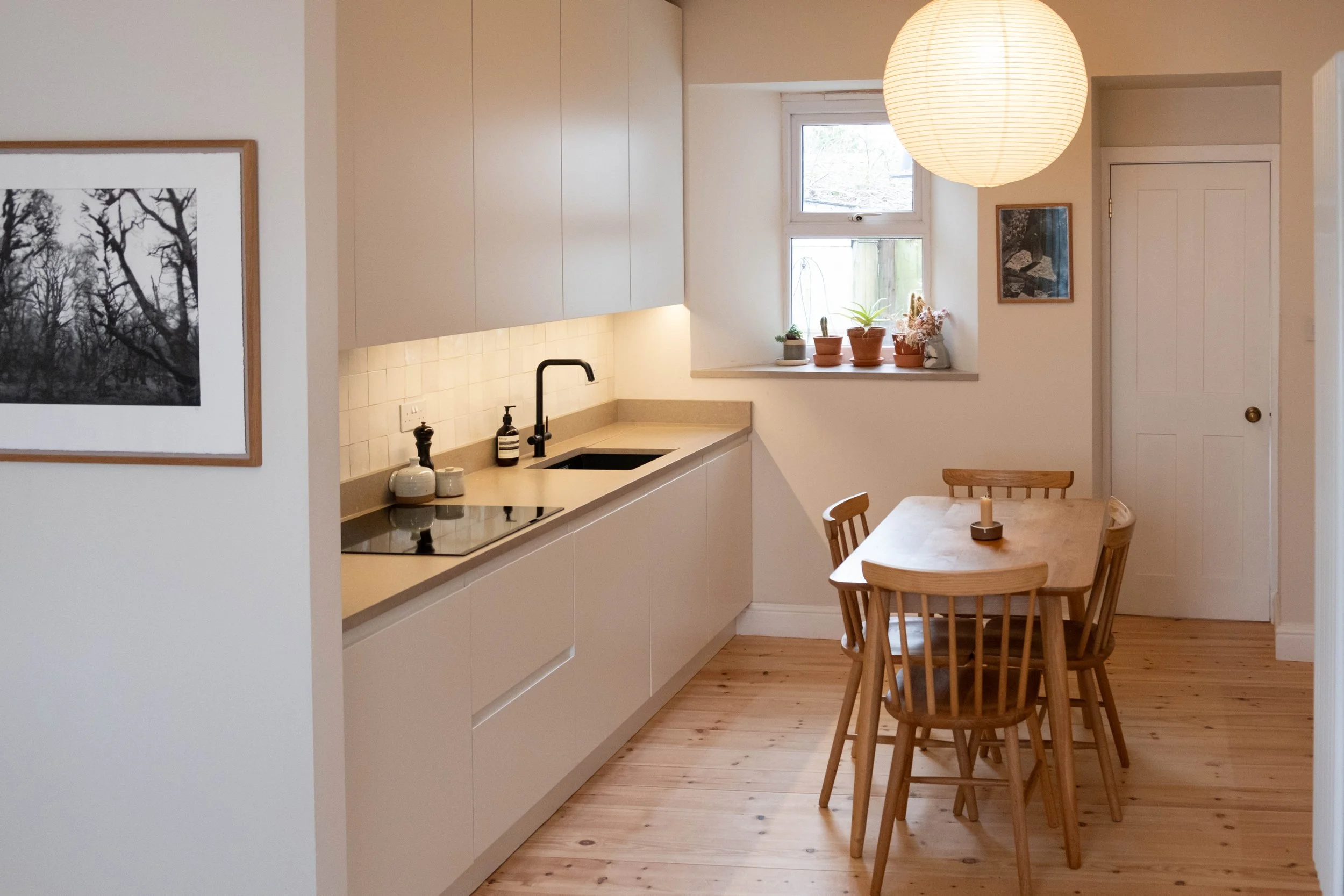 A cozy kitchen with white cabinets, a beige countertop, a black sink and faucet, a small dining table surrounded by four wooden chairs, windows with small potted plants on the sill, a framed black-and-white photograph on the wall, and a white paper l