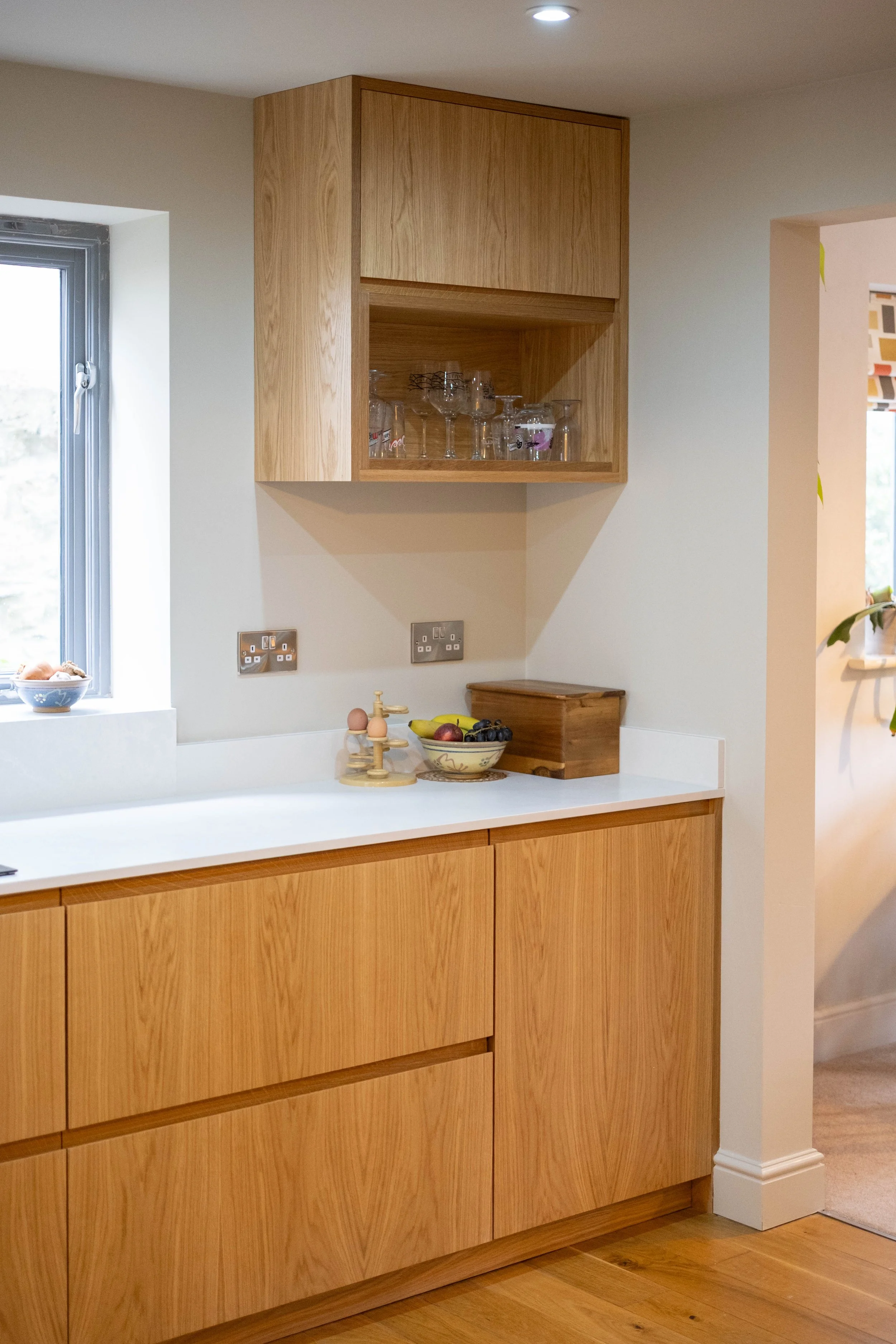 A wooden kitchen cabinet with a closed upper compartment and open shelf in a modern kitchen. Glassware is stored on the open shelf. Below the cabinet, there is a white countertop with a fruit bowl and a wooden cutting board. The kitchen has a window 