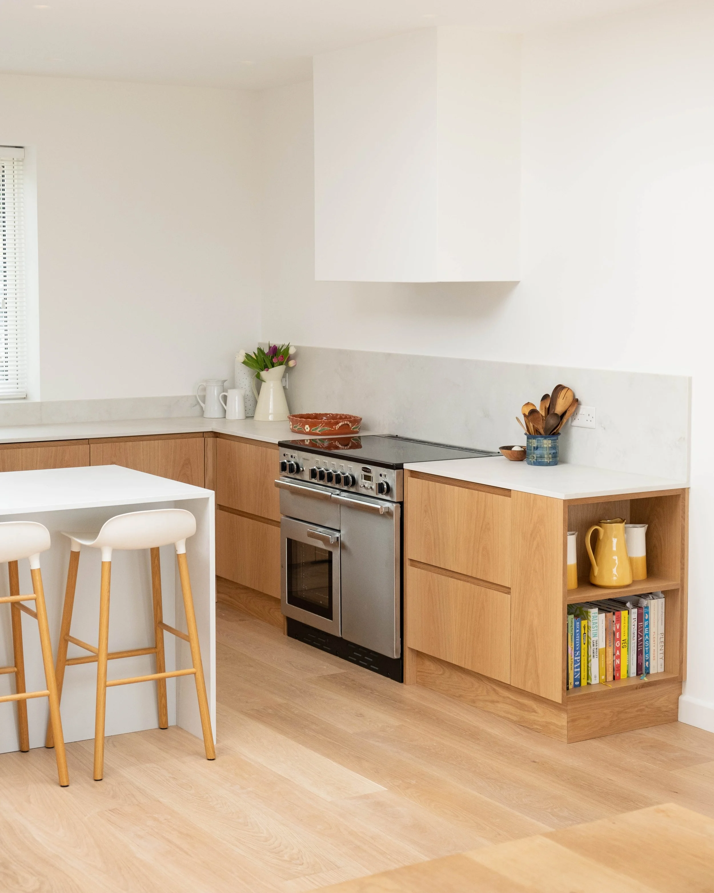 A modern kitchen with light wood cabinets, a stainless steel stove, a white countertop, and various decorative items including a vase with flowers, a terracotta bowl, and a blue container holding wooden utensils. There is also a bookshelf with colorf