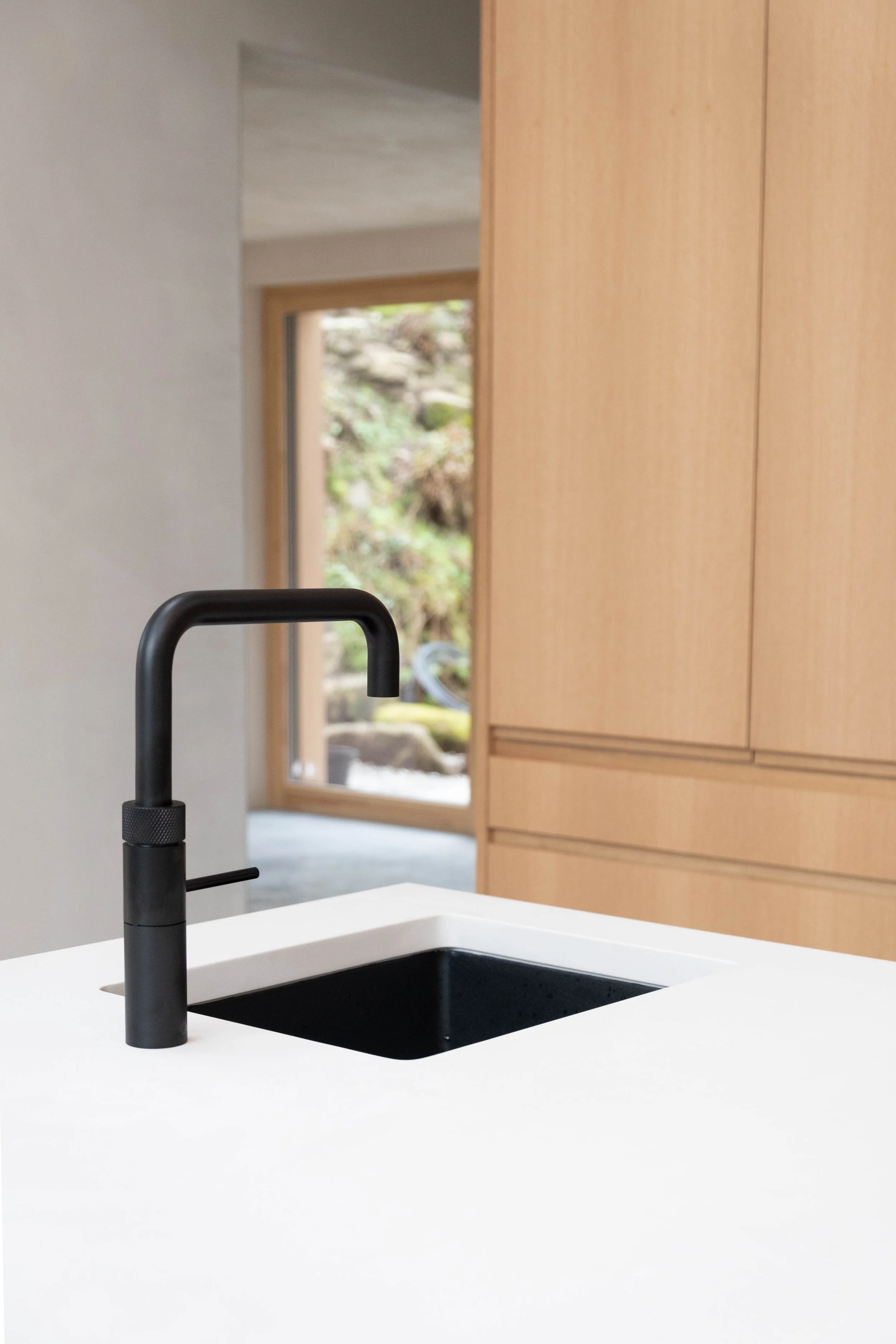 Modern kitchen with black faucet, white sink, wooden cabinets, and a window showing a garden outside.