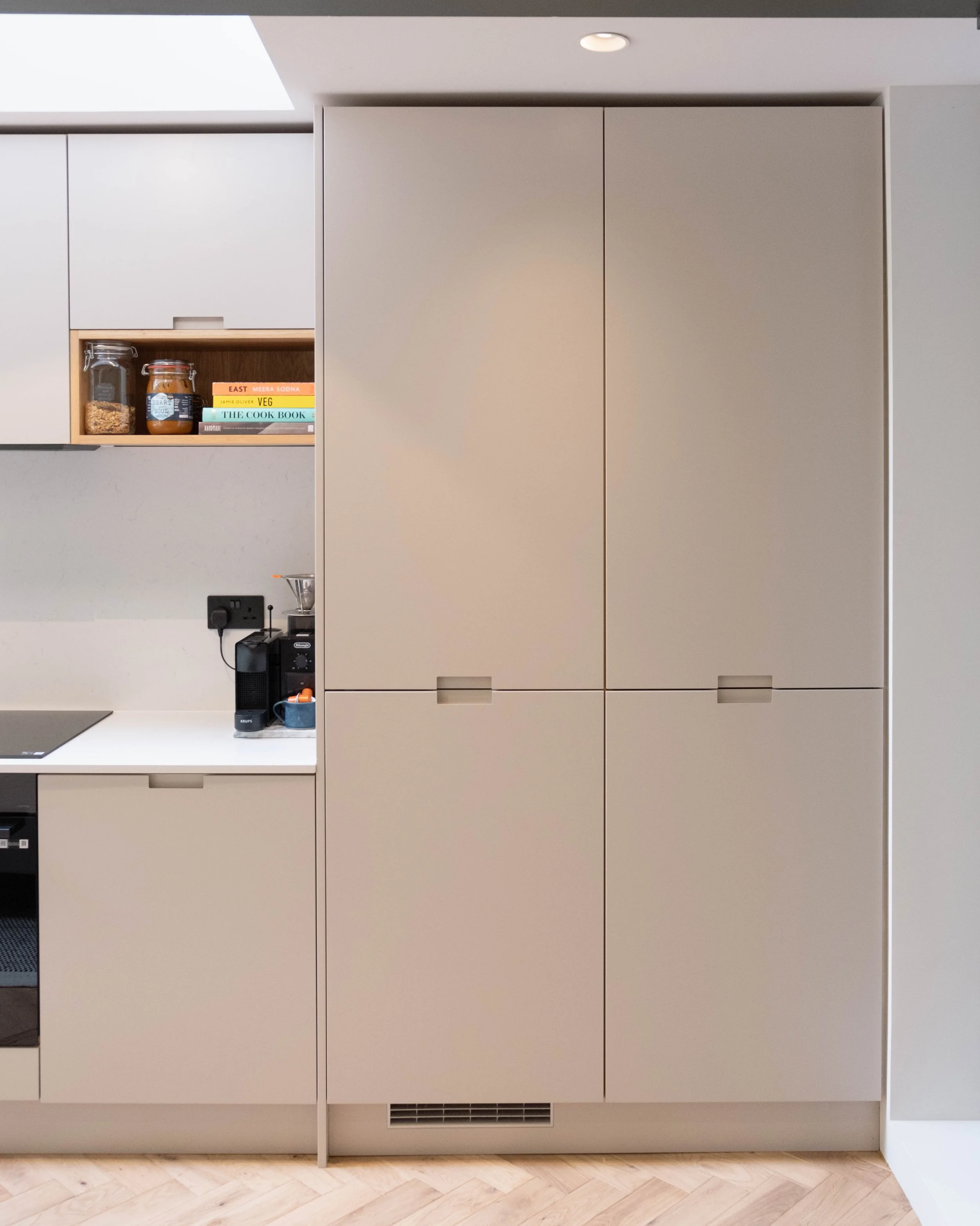 A modern kitchen cabinet with closed doors, beige in color, located against a wall painted in a light neutral shade. There is a small open shelf on the left side of the cabinet showing jars, a cookbook, and decorative items. A potted plant, a black p