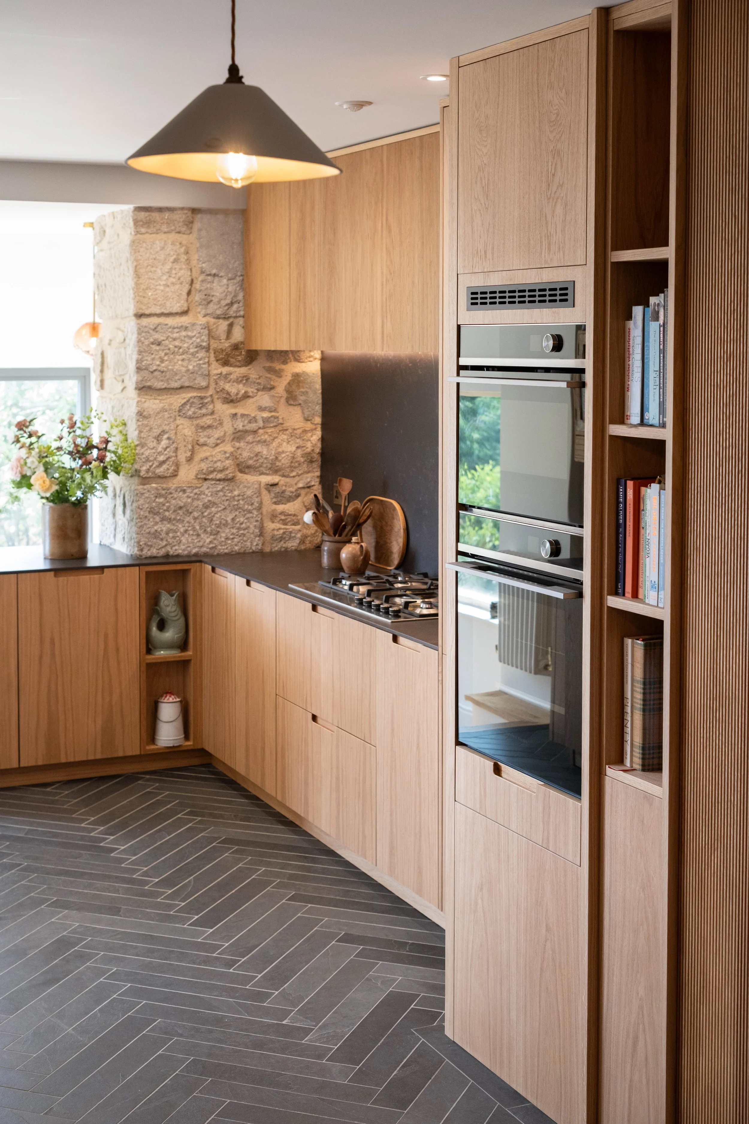 Modern kitchen with wooden cabinets, stone wall feature, black countertops, and gray herringbone floor tiles. Includes built-in oven, shelves with books, and a vase with flowers.