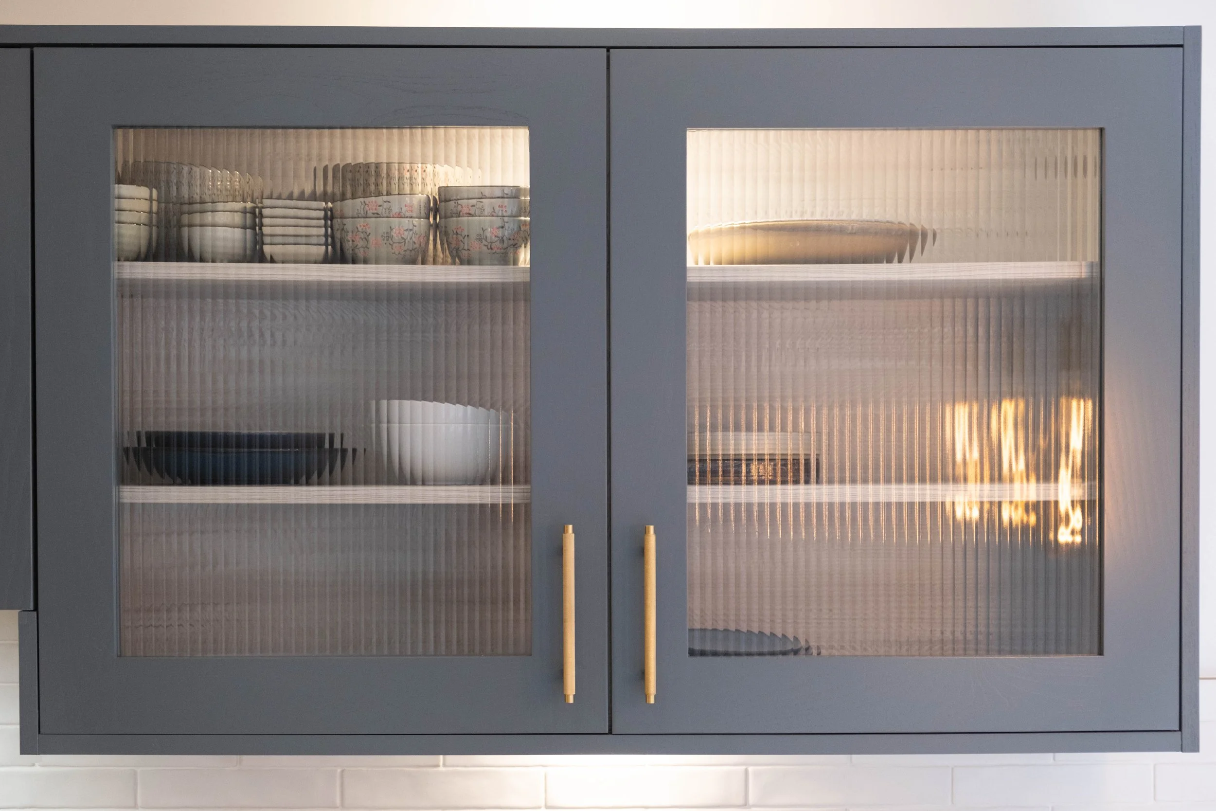 Kitchen cabinet with glass doors containing stacked dishes and bowls inside, illuminated from within.
