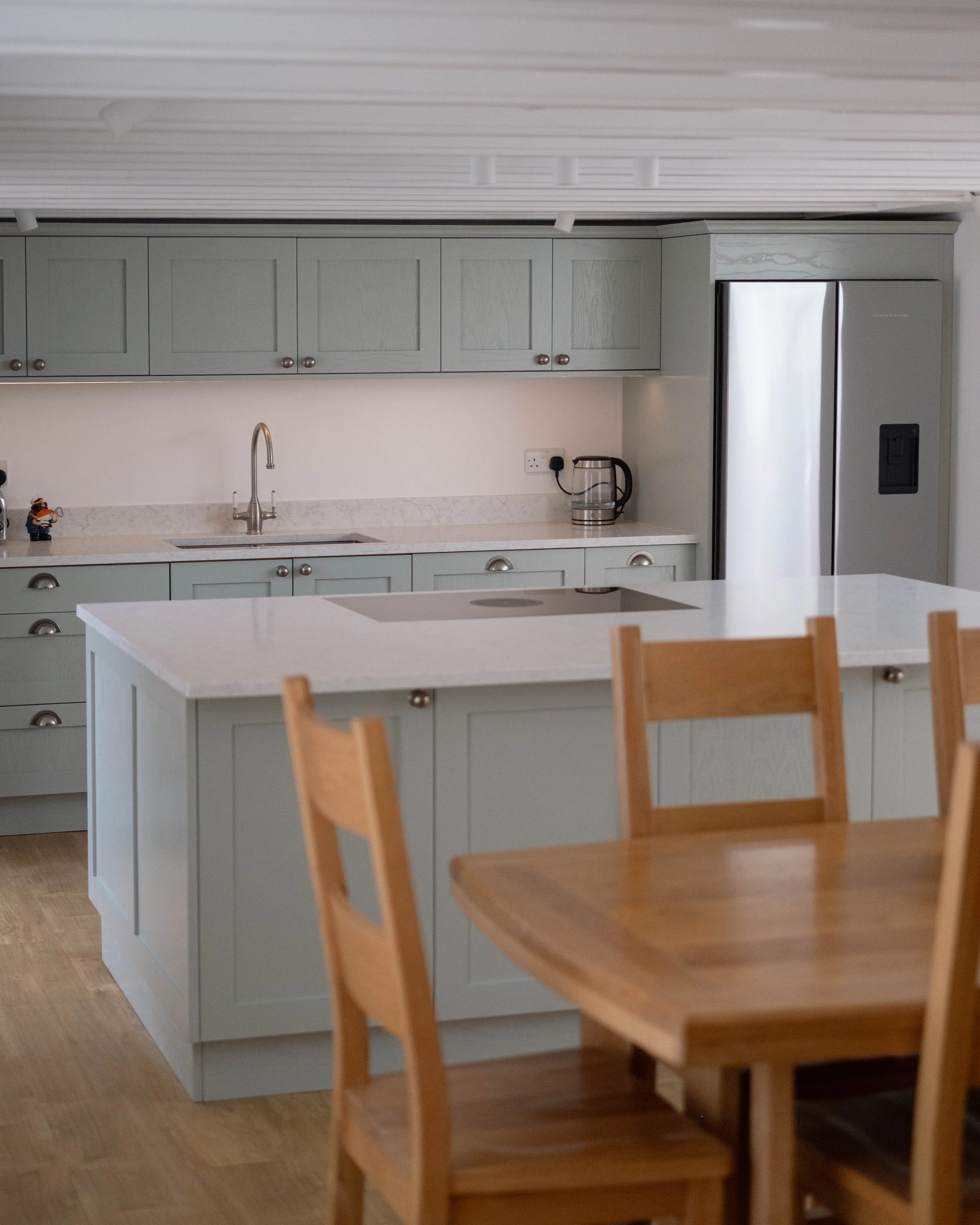 A kitchen with light gray cabinets, a white countertop, a stainless steel refrigerator, and a wooden dining chair in the foreground.