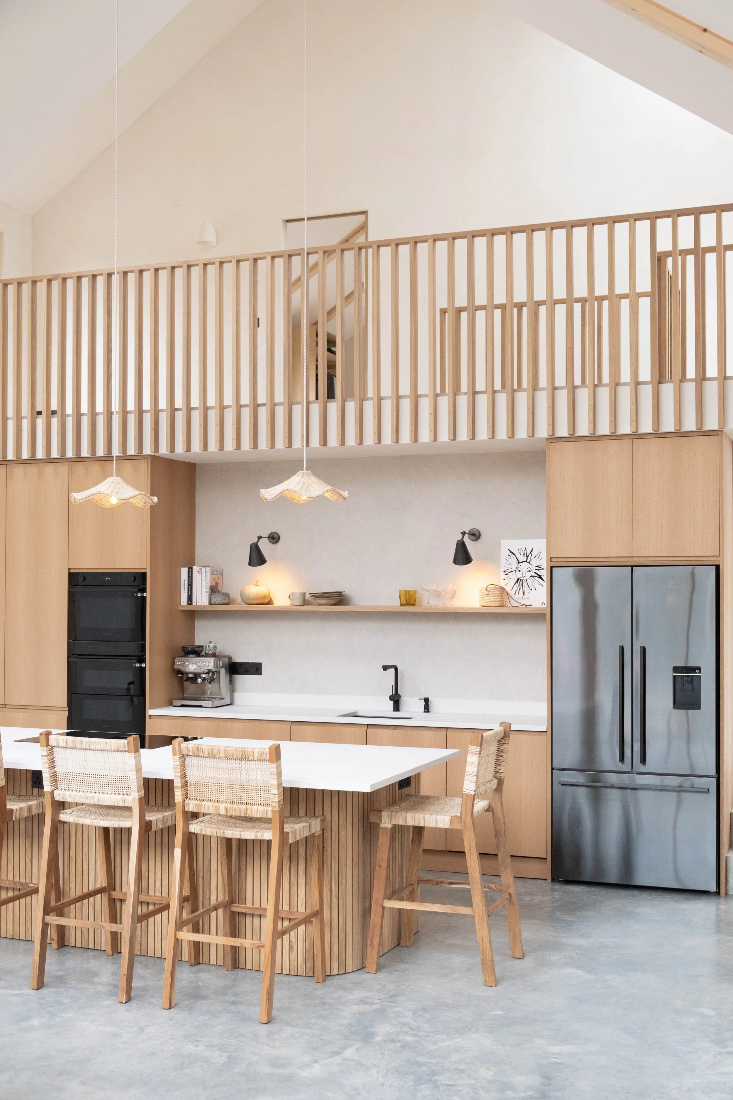 Modern kitchen with light wood cabinetry, a white countertop, black faucet, stainless steel refrigerator, and a dining table with wooden chairs. There is an open loft area with a wooden railing above the kitchen.