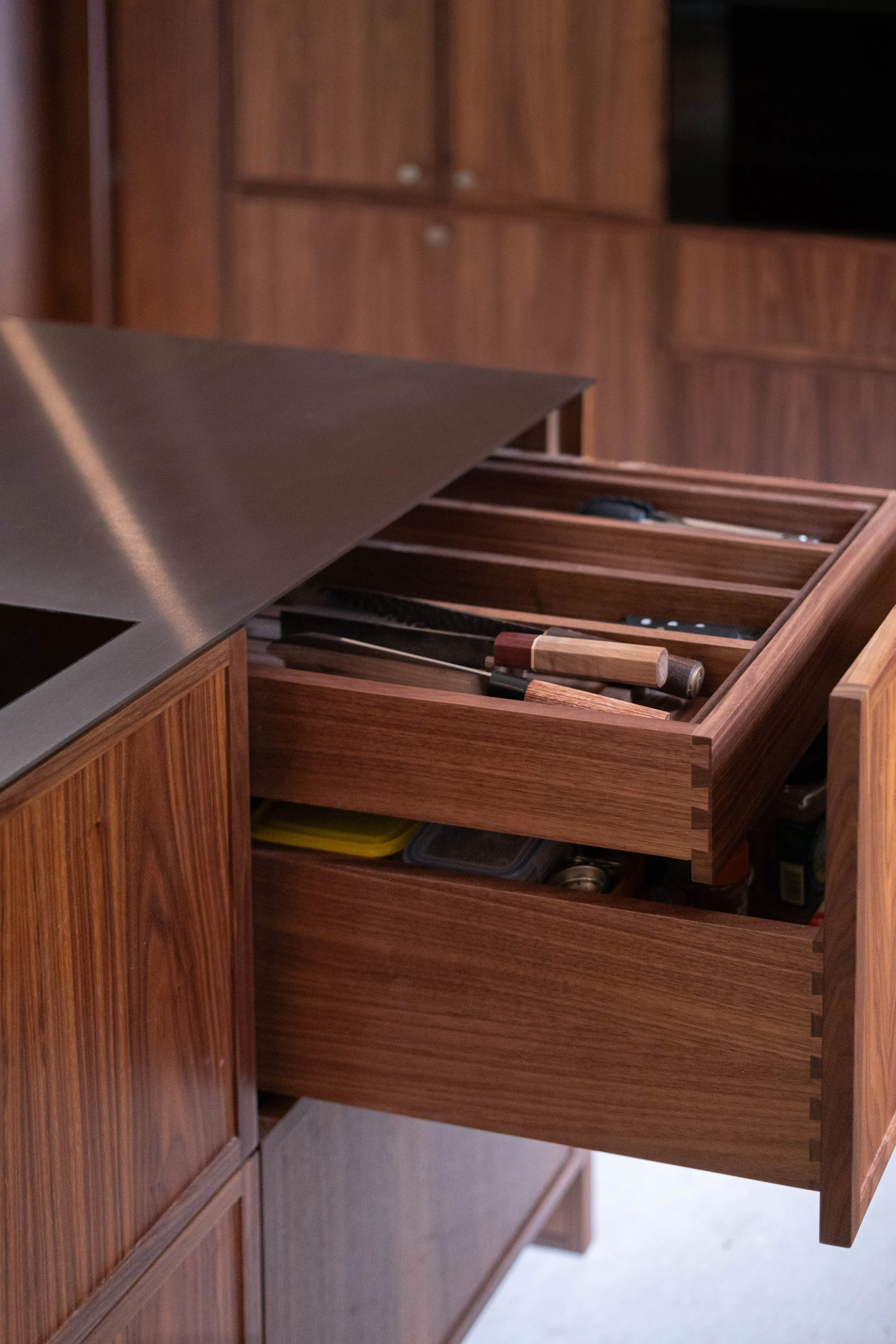 Open wooden kitchen drawer filled with knives and cooking utensils, against a background of wooden cabinet doors.