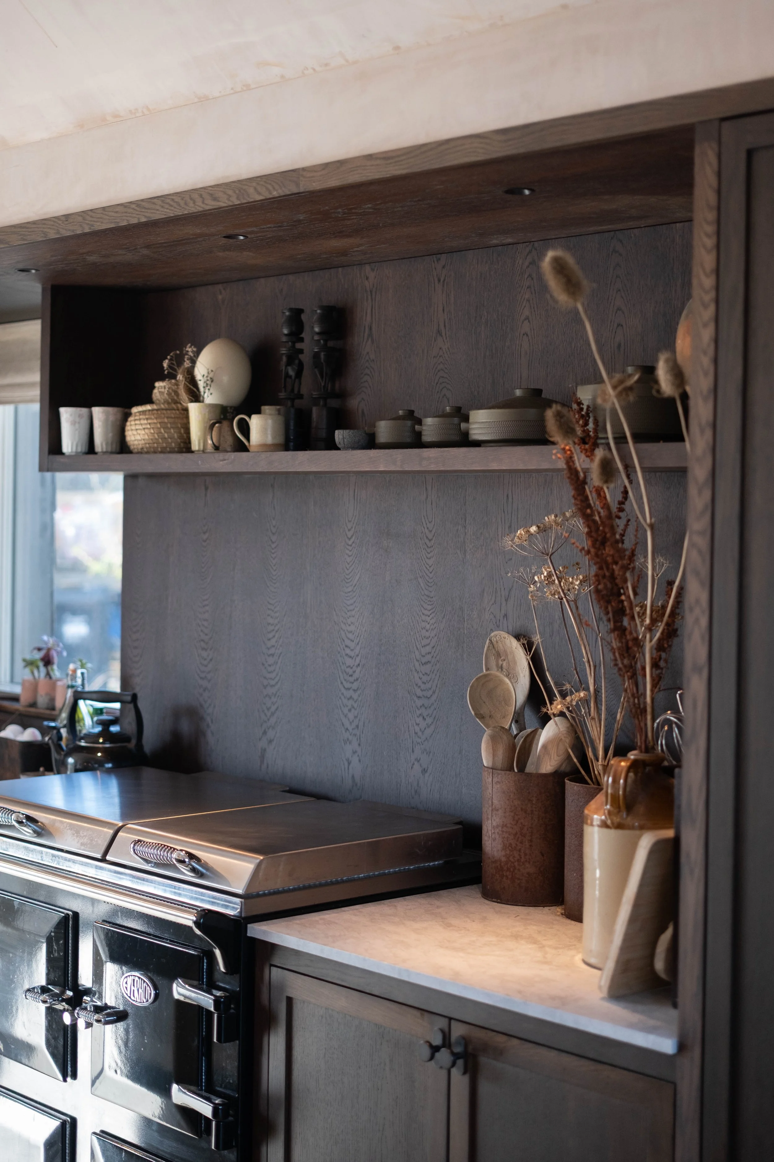 A kitchen with dark wood cabinets, a stainless steel stove, and decorative items including vases with dried flowers on the countertop, and a dark wood shelf with ceramic dishes, cups, and trays.
