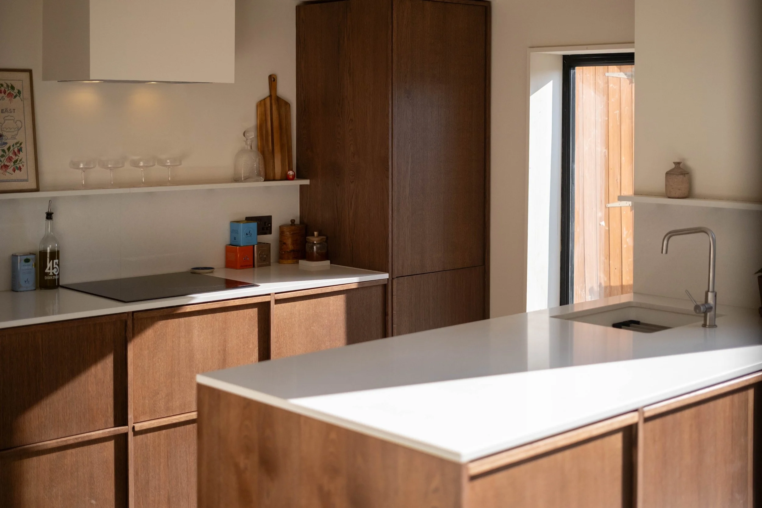 A modern kitchen with wooden cabinets and white countertops. There is a sink with a silver faucet, a black stove, and shelves with decorative items, glassware, and bottles. A door leads outside, revealing a wooden fence.