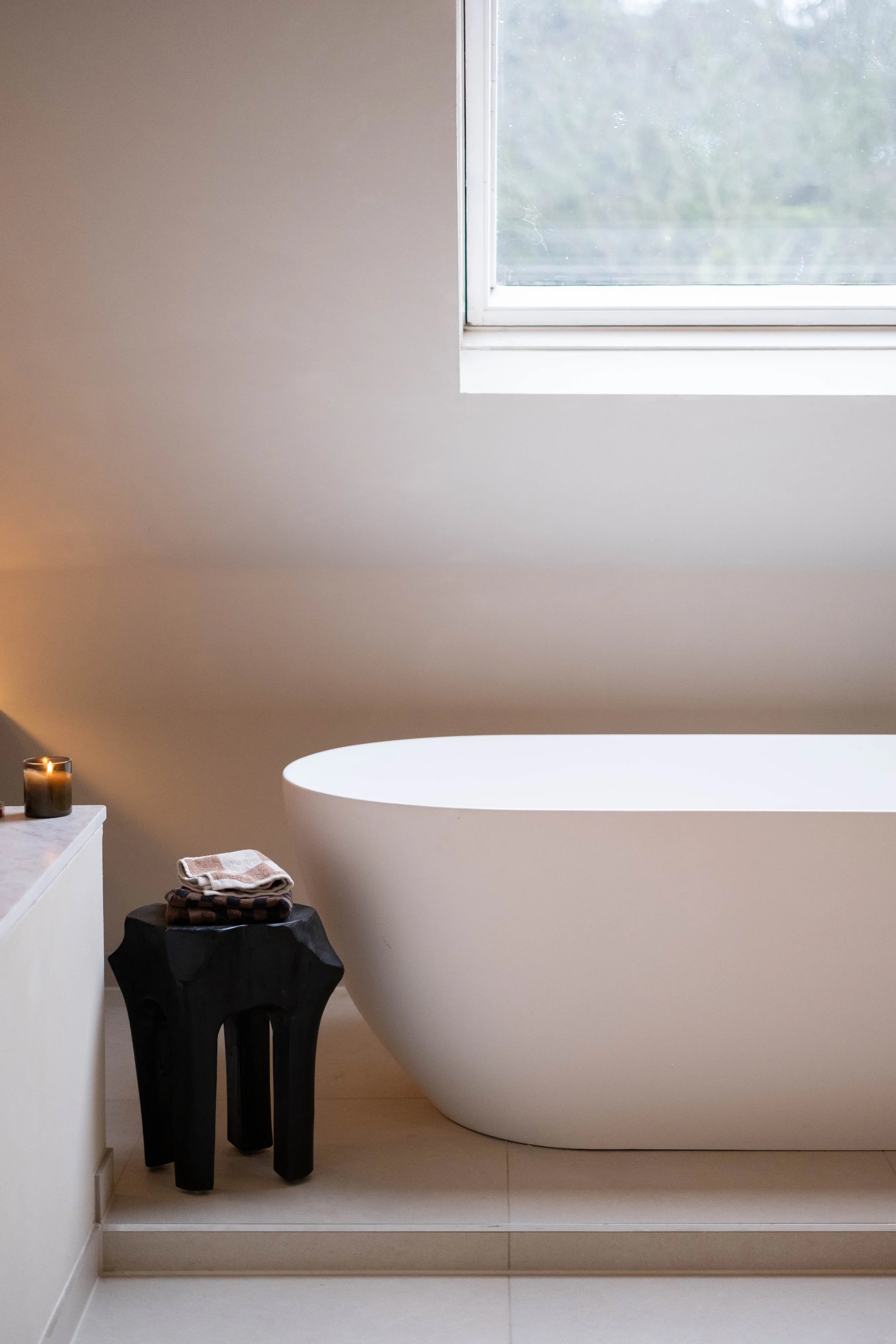 A bathroom with a white bathtub, a small black side table with a folded towel on top, a lit candle on the counter, a white cabinet, and a window with a view of blurry greenery outside.