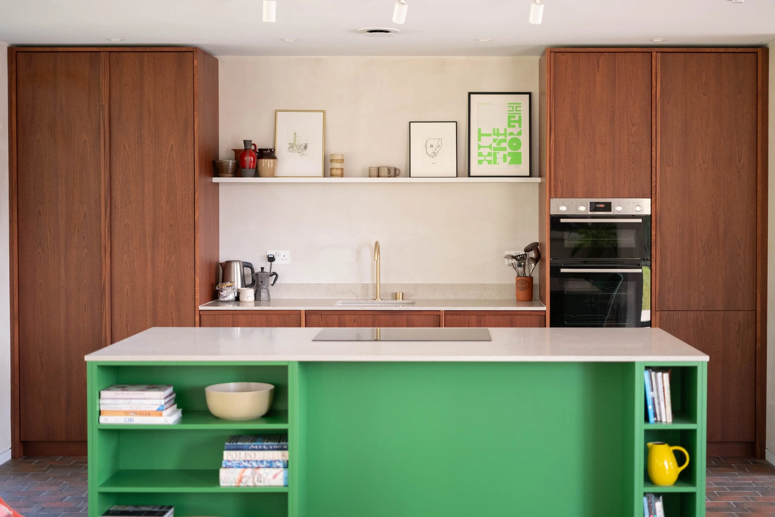 Modern kitchen with wooden cabinetry, a white countertop, and a vibrant green island. Decor includes framed art, vases, a kettle, and cook utensils. Stainless steel oven built into the cabinetry, and a yellow pitcher on the floor.
