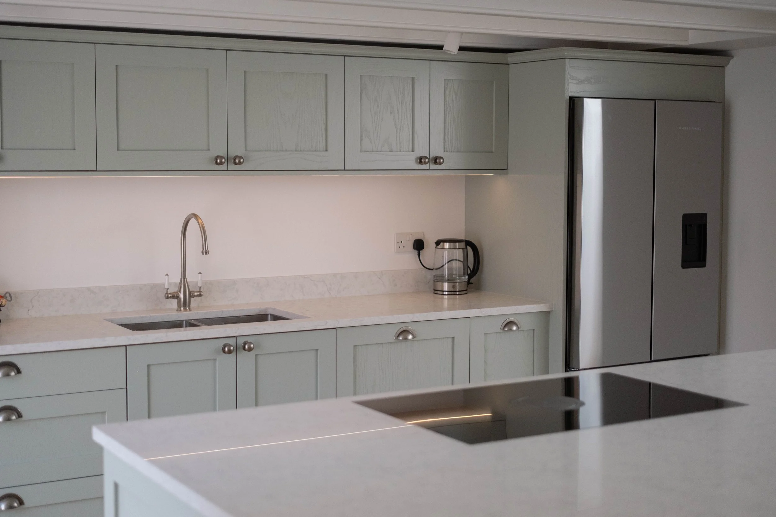 A modern kitchen with light green cabinets, marble countertops, a stainless steel refrigerator, a sink with a silver faucet, and a glass electric kettle on the counter.