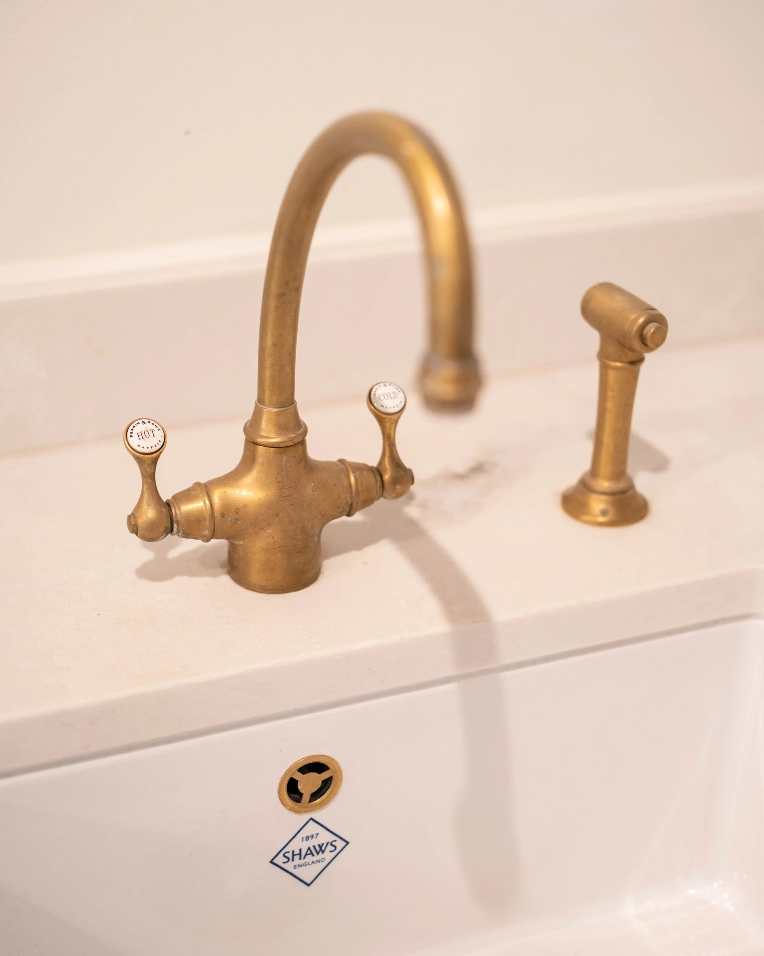 Close-up of a vintage brass faucet with two handles labeled 'HOT' and 'COLD', mounted on a white porcelain sink with a 'Shaws England' logo sticker visible.