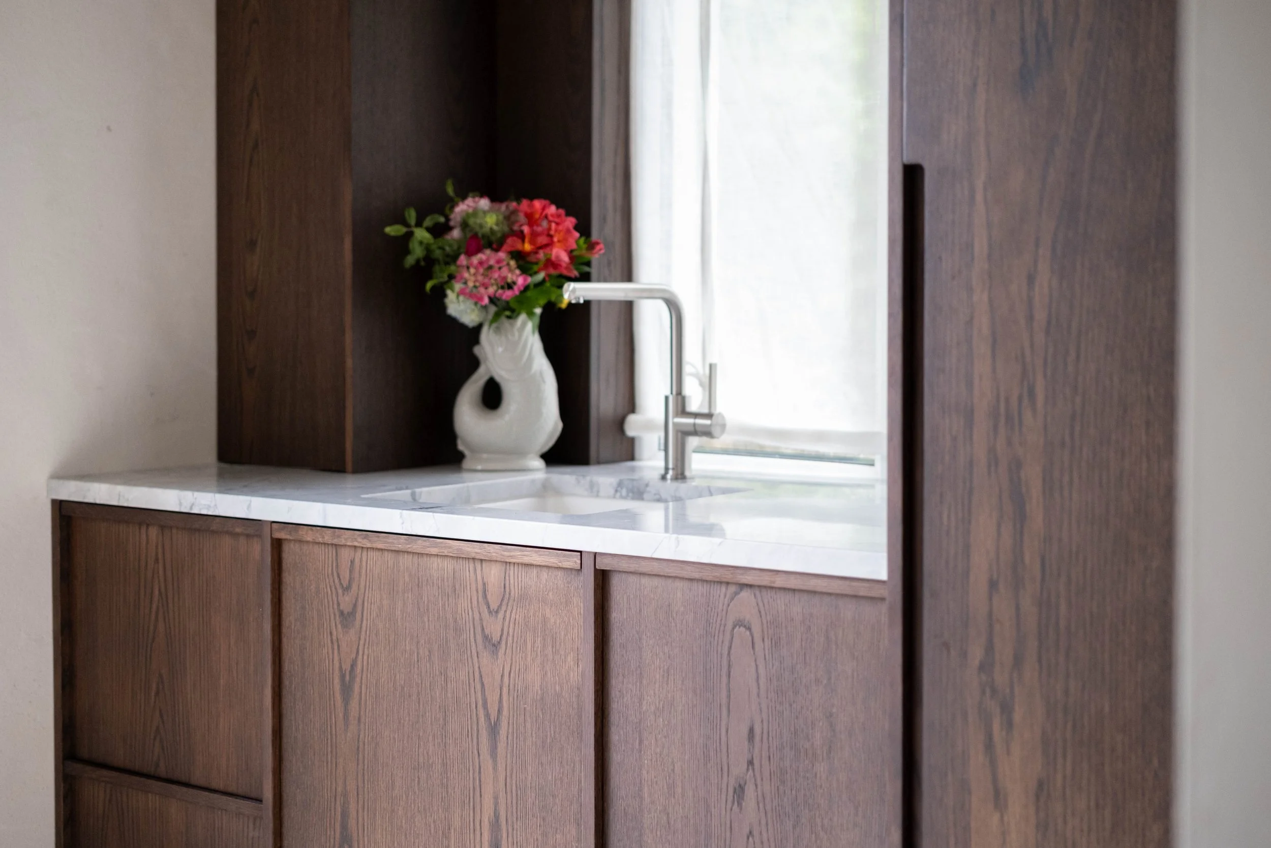 A kitchen sink with a white marble countertop, a modern silver faucet, and a dark wooden cabinet. A white vase with colorful pink, red, and orange flowers is positioned on the countertop near the faucet. There is a window with sheer white curtains in the background.