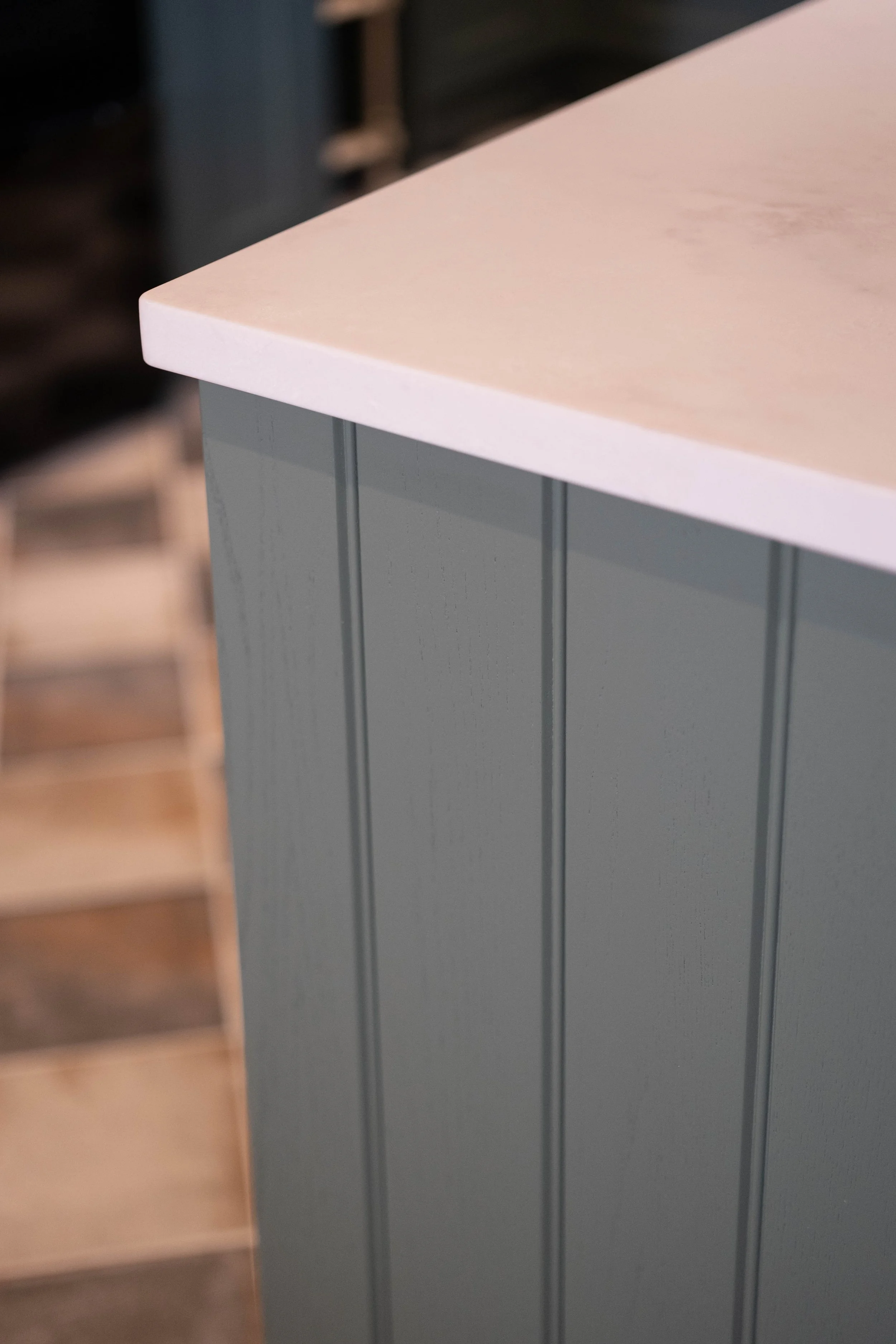 Close-up of a partial view of a white countertop and a gray cabinet with vertical lines, with a blurred background showing a wooden staircase.