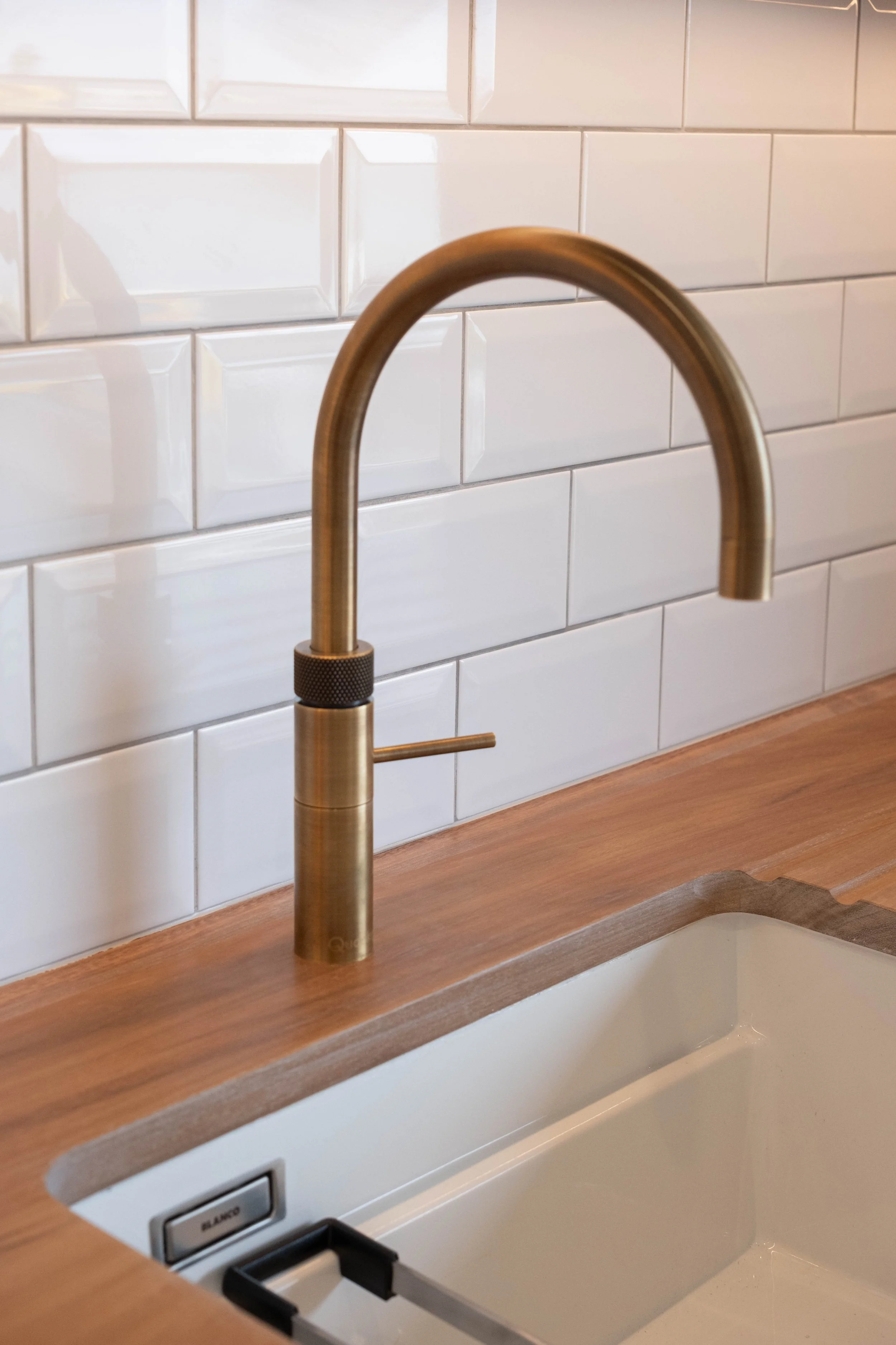 A kitchen sink with a modern, curved, brushed metal faucet on a wooden countertop, white subway tile backsplash, and an electrical outlet below.