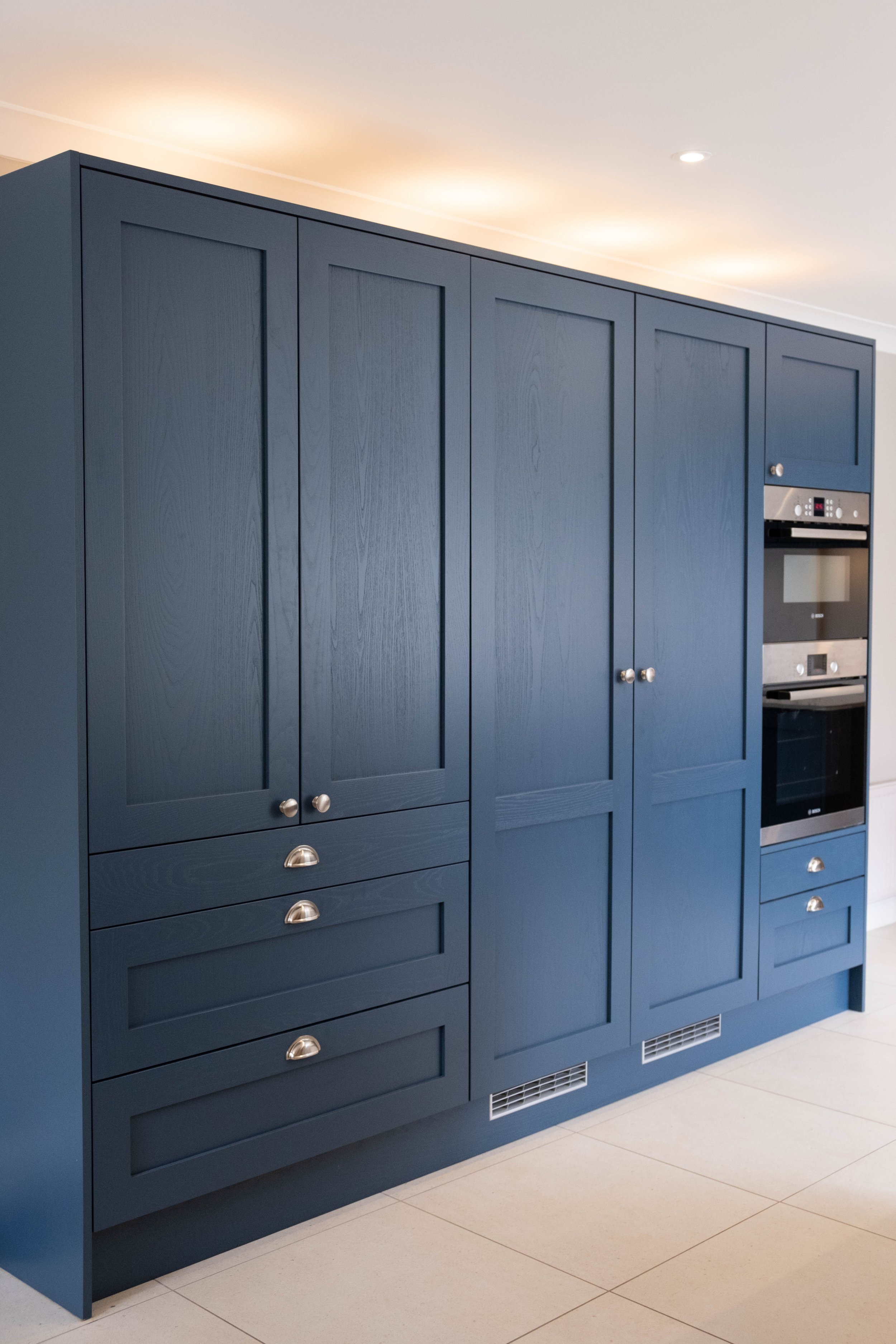 Blue kitchen cabinets with silver knobs and drawers, built-in oven, white tiled floor, ceiling with recessed lighting.