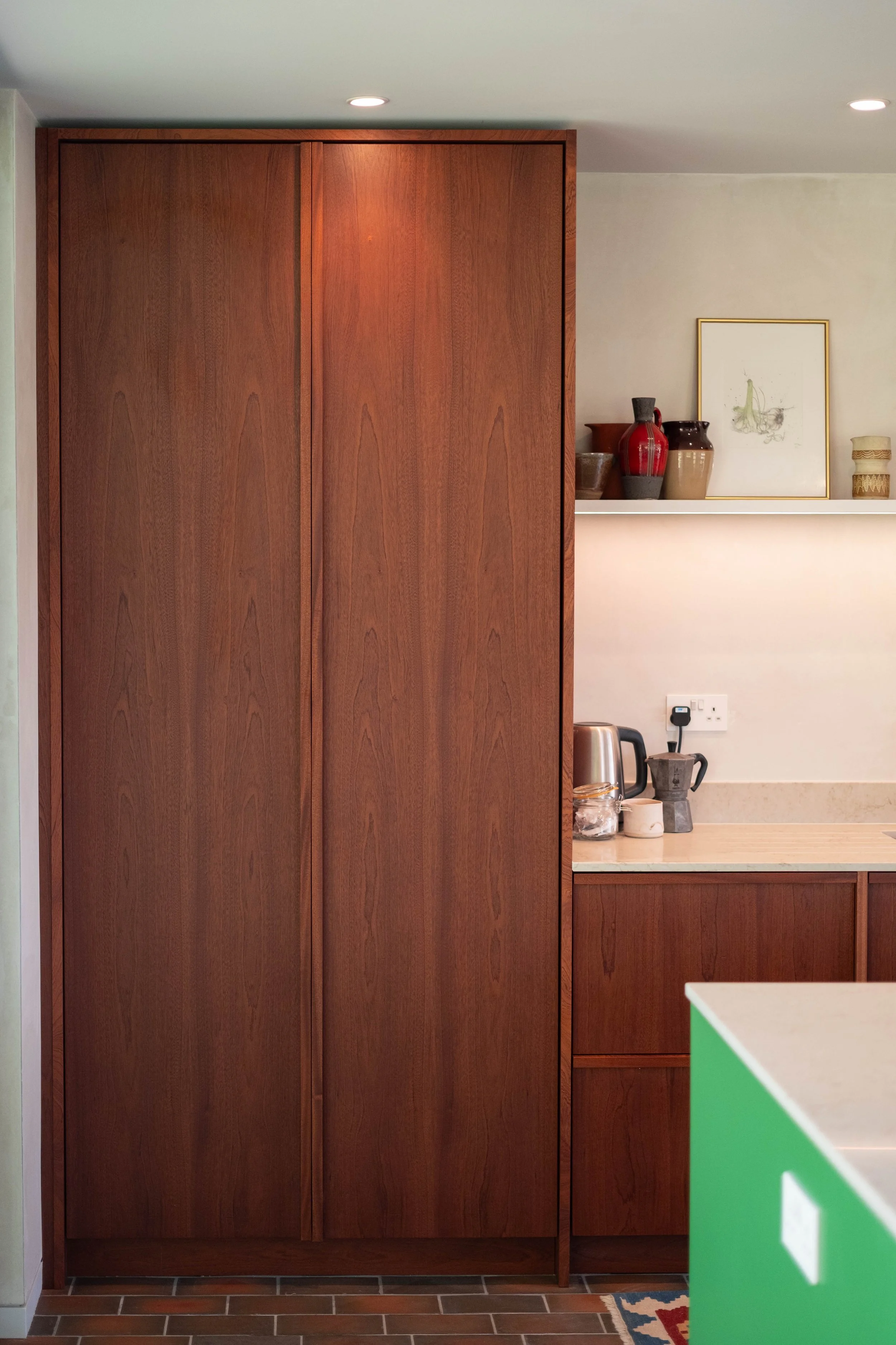 Interior view of a wooden kitchen or living space with a large wooden cabinet, a shelf with decorative vases, a framed artwork, and a countertop with a kettle, a moka pot, and other items.