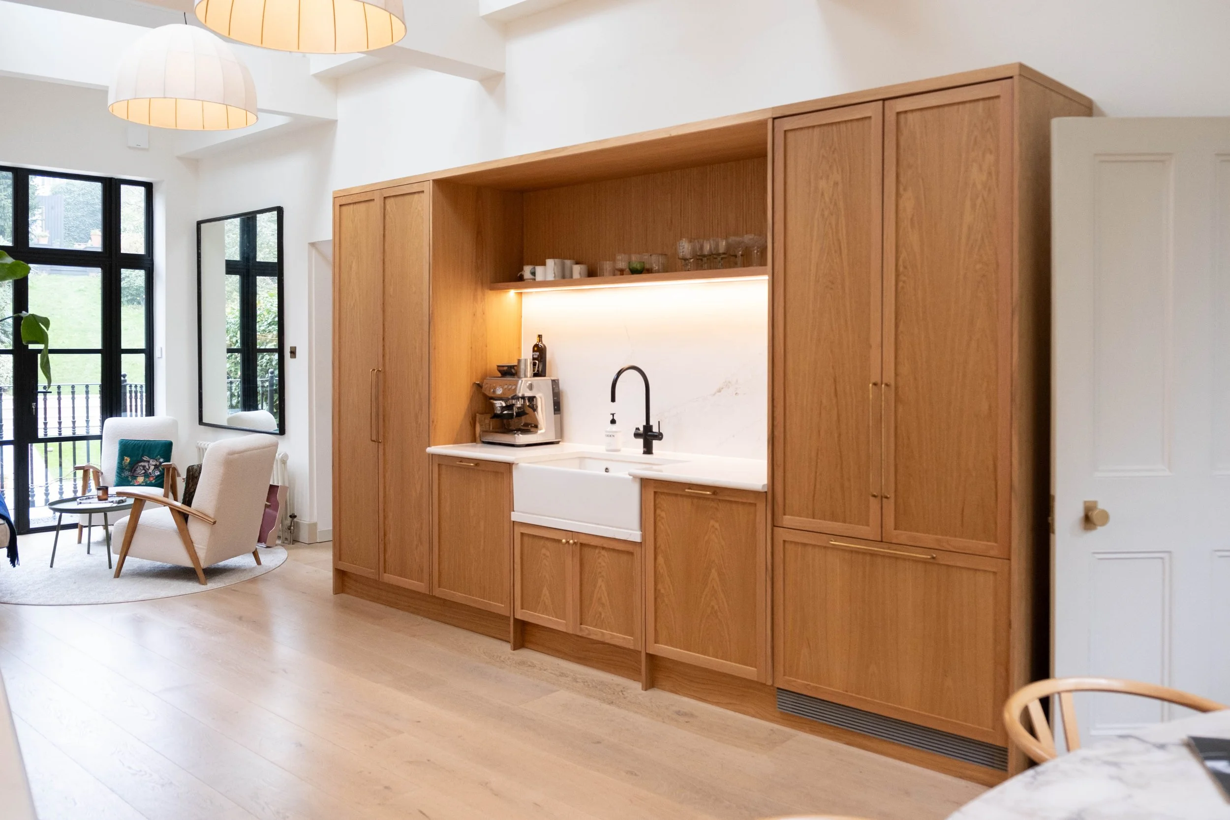 Interior view of a modern kitchen with wooden cabinetry, a white farmhouse sink, black faucet, and a coffee machine on the countertop. The room has large windows and natural light, with a cozy seating area nearby.