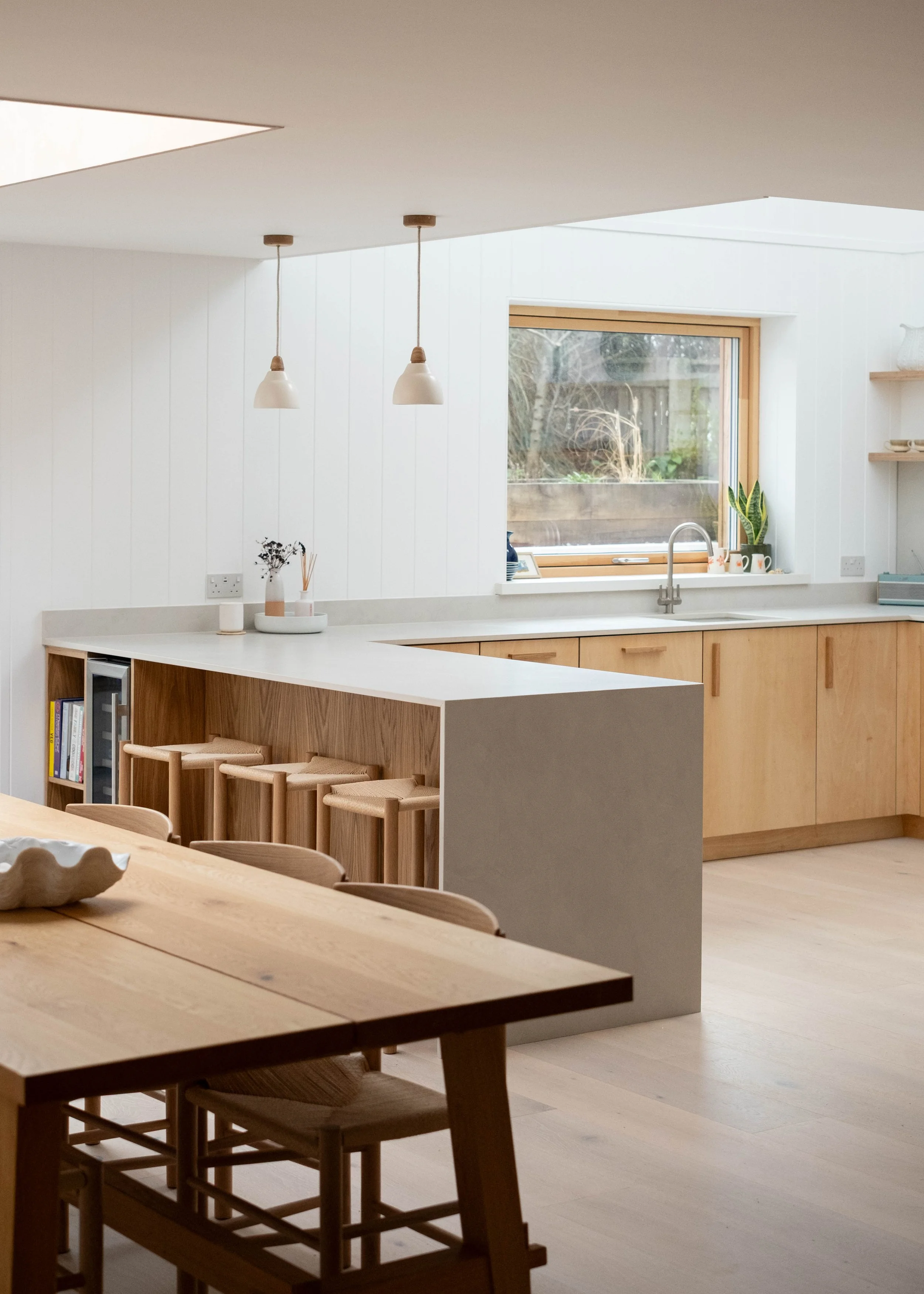 A modern kitchen and dining area with natural wood cabinets, white countertops, and pendant lights hanging from the ceiling. There is a window with a view of a garden, and a potted plant on the windowsill.