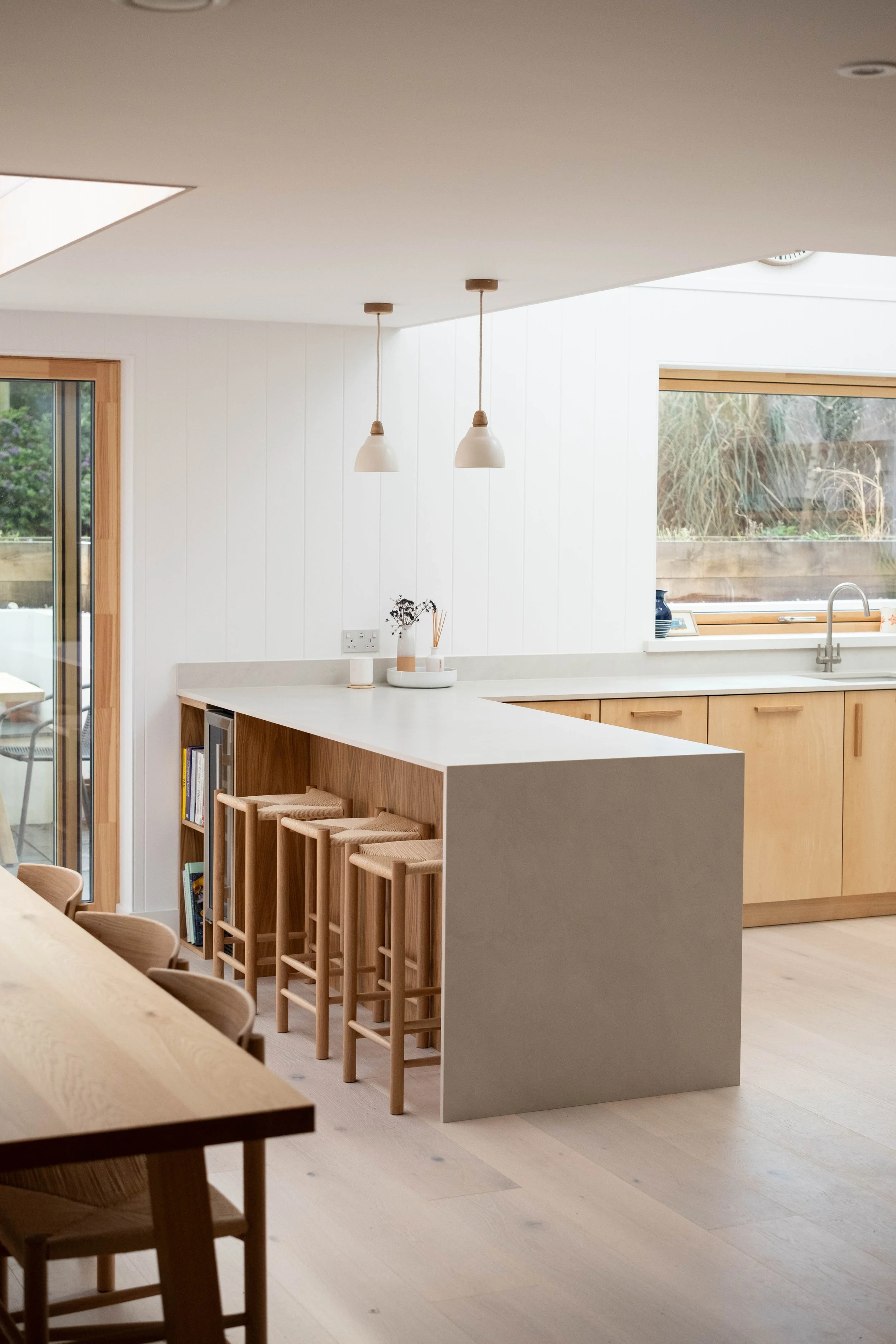 Modern minimalist kitchen with white walls, wooden cabinets, large window with outdoor view, and a white kitchen island with seating.