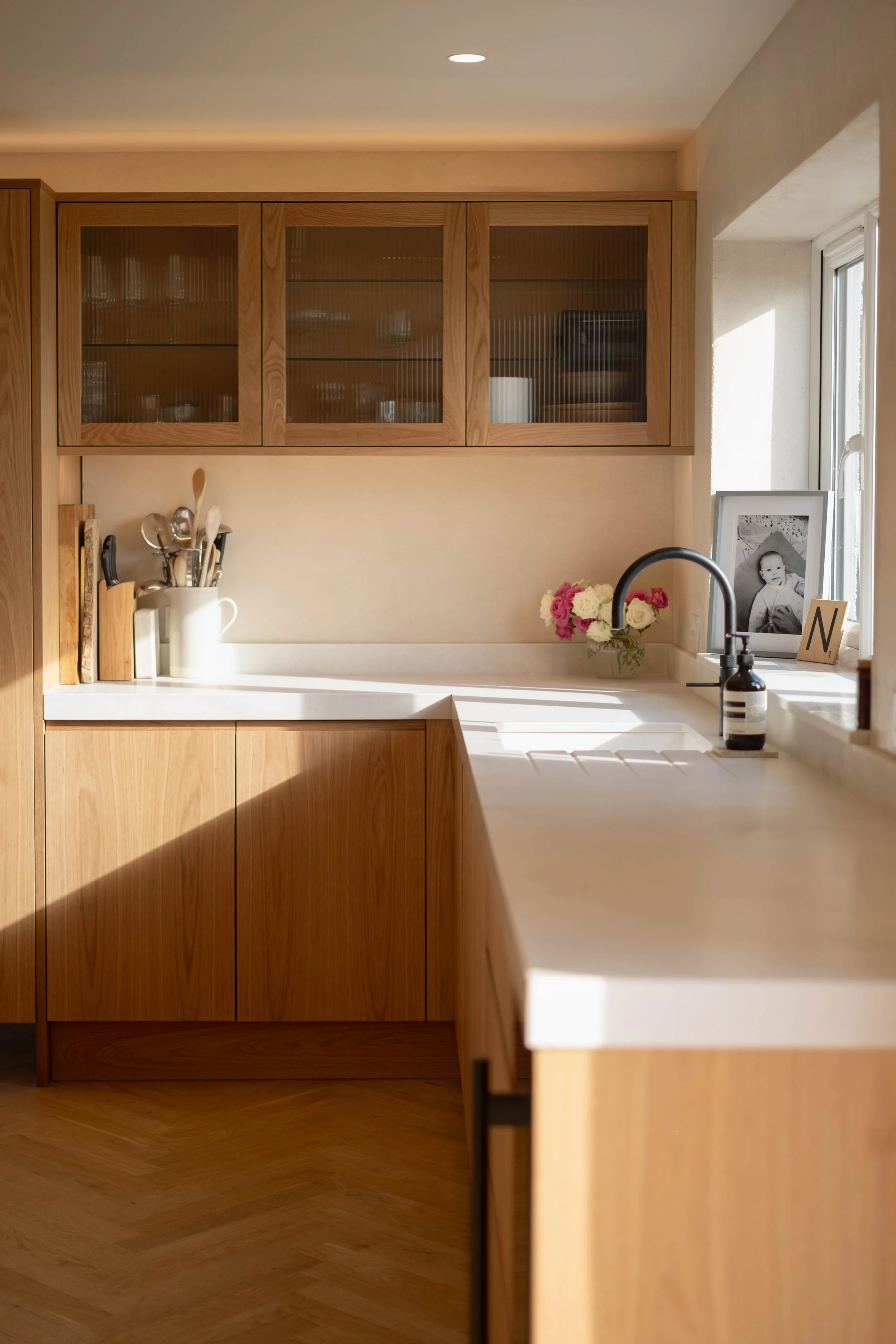 A kitchen countertop with a black faucet, a photo of a baby in a white frame, a bouquet of pink and white flowers, a soap dispenser, a white pitcher holding utensils, and a window with sunlight streaming in.