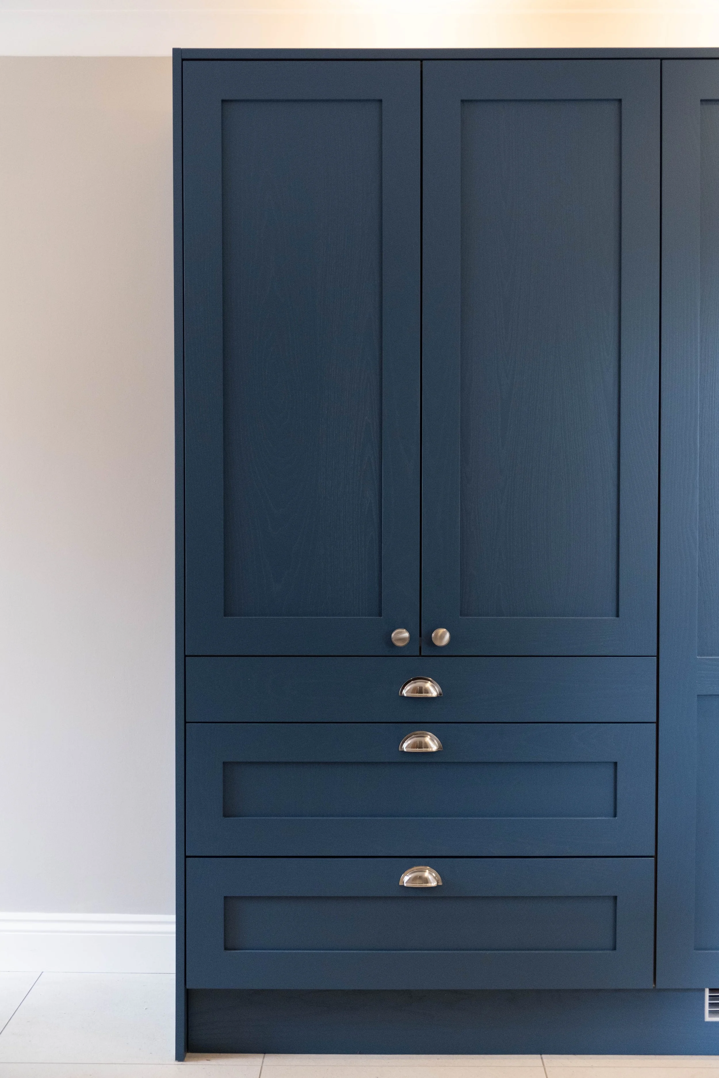 A tall, dark blue kitchen cabinet with three drawers at the bottom and double doors above. The cabinet has metallic, semi-circular handles. It is positioned against a light-colored wall with white tile flooring.