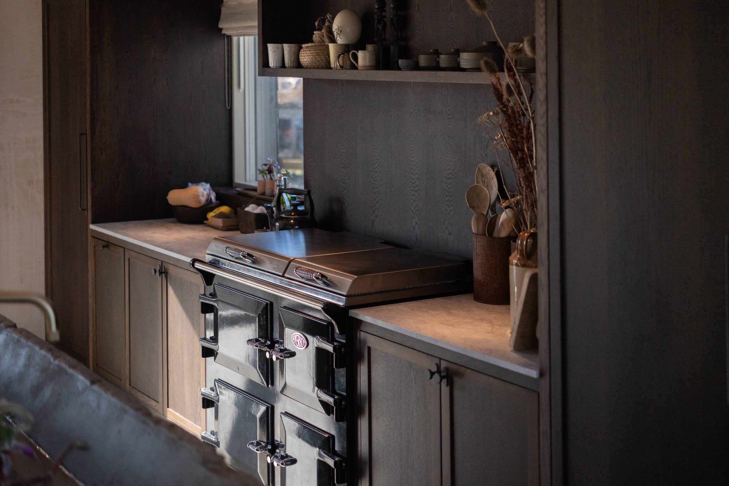 Cozy kitchen with dark wooden cabinets, a vintage steel oven, and a marble countertop. Open shelves display cups, bowls, and decorative items. A window lets in natural light, and dried flowers sit in a vase.