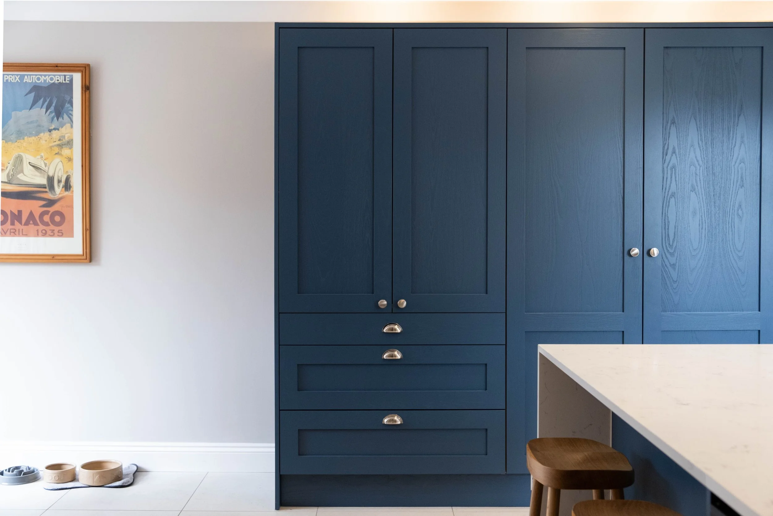 A modern kitchen corner with a large blue cabinet with silver knobs, a white countertop with wooden stools, a white wall with a vintage automotive poster, and some pet bowls on the floor.