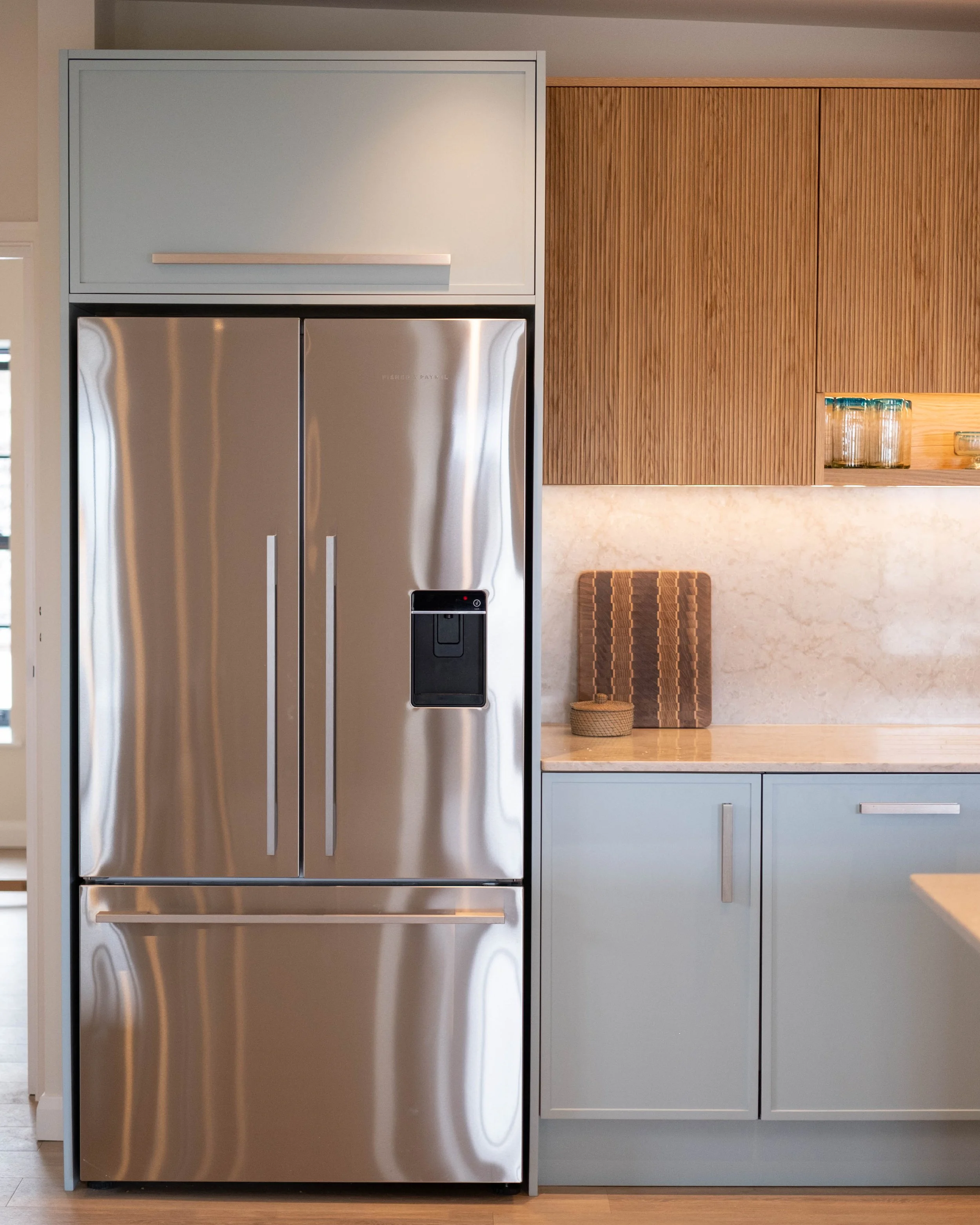 Modern kitchen with stainless steel refrigerator, light blue cabinets, beige wall, and wooden paneling with glass jars on a shelf.