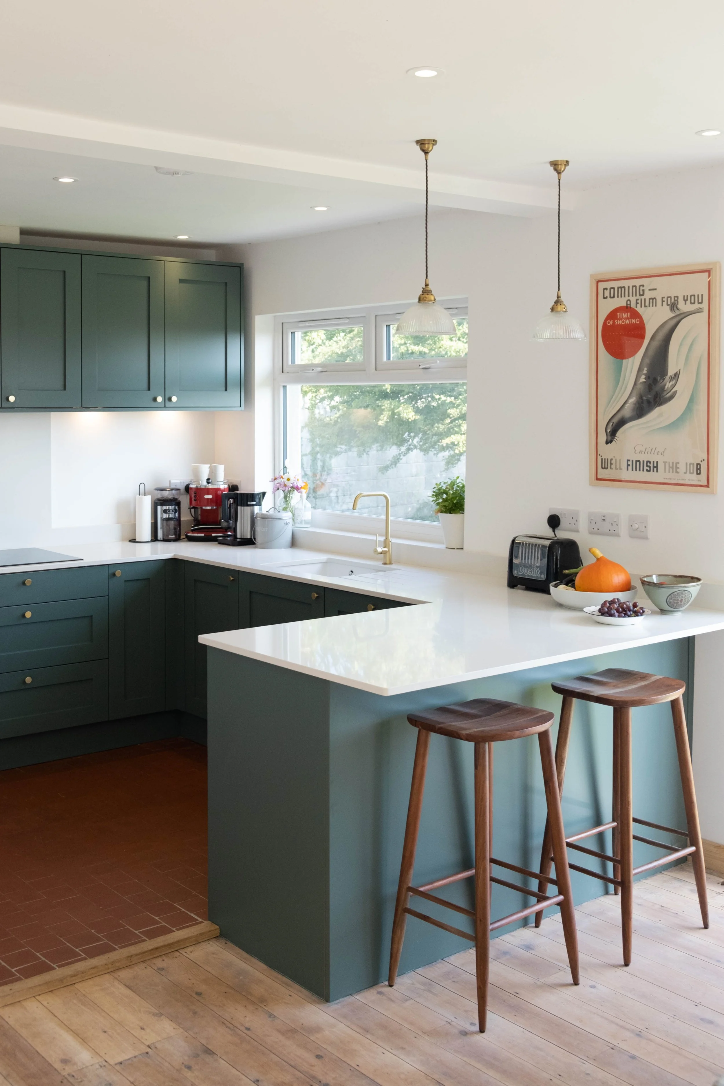Kitchen with green cabinets, white countertop, two wooden bar stools, hanging pendant lights, window with greenery outside, and a wall poster.