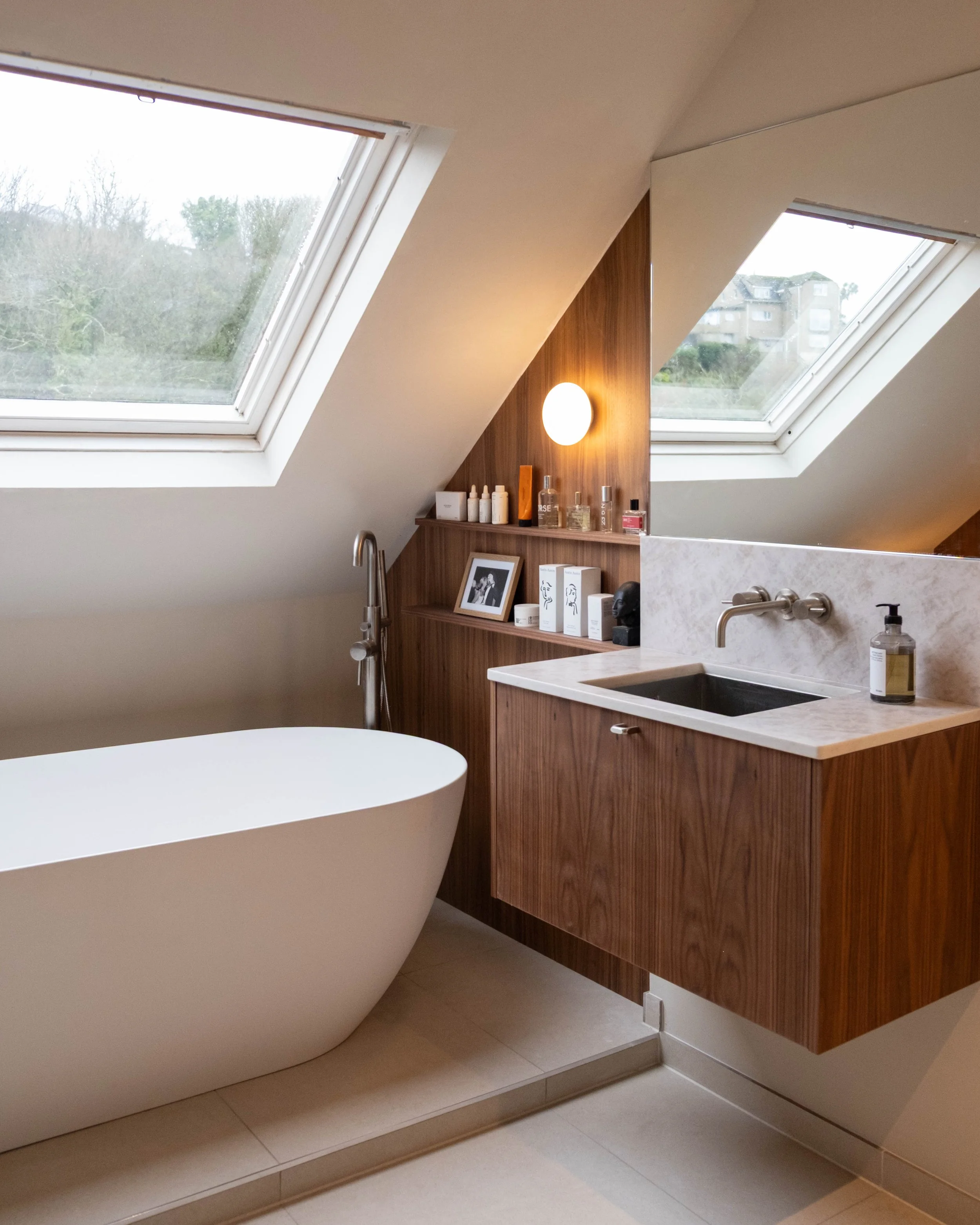 Corner of a modern bathroom with skylight windows, a wooden cabinet with bottles and framed photos, and a white bathtub.