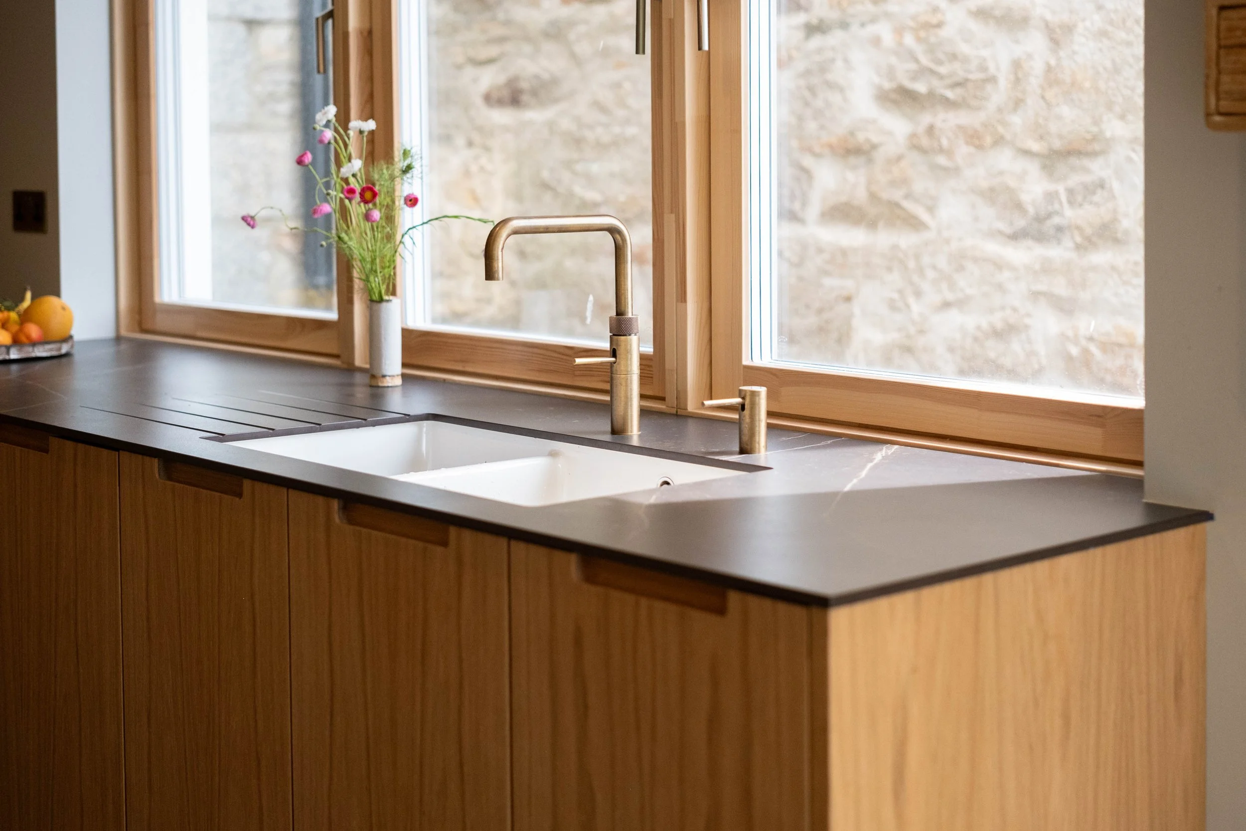 A kitchen sink with a brass faucet, placed under a large window with a view of a stone wall outside. There is a vase with flowers and a fruit bowl on the dark countertop.