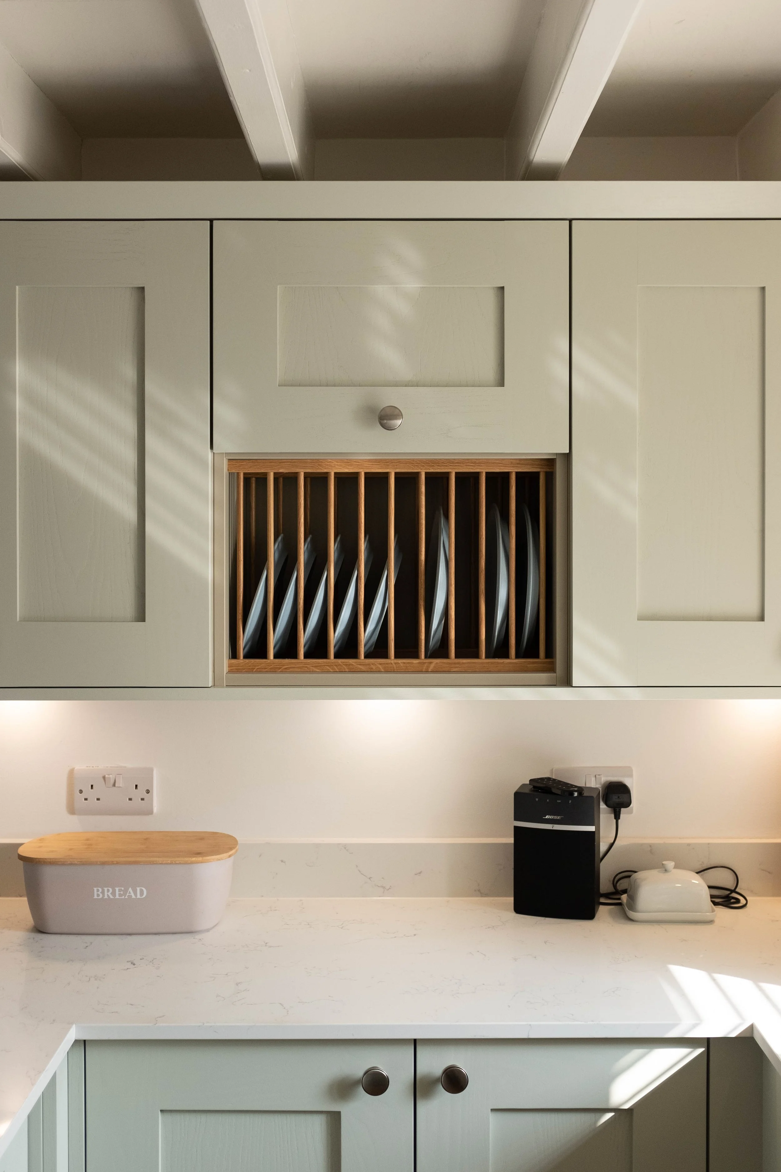 Kitchen countertop with a beige bread box labeled 'BREAD', a black speaker, a white kettle, and a white wall with electrical outlets and a black switch.
