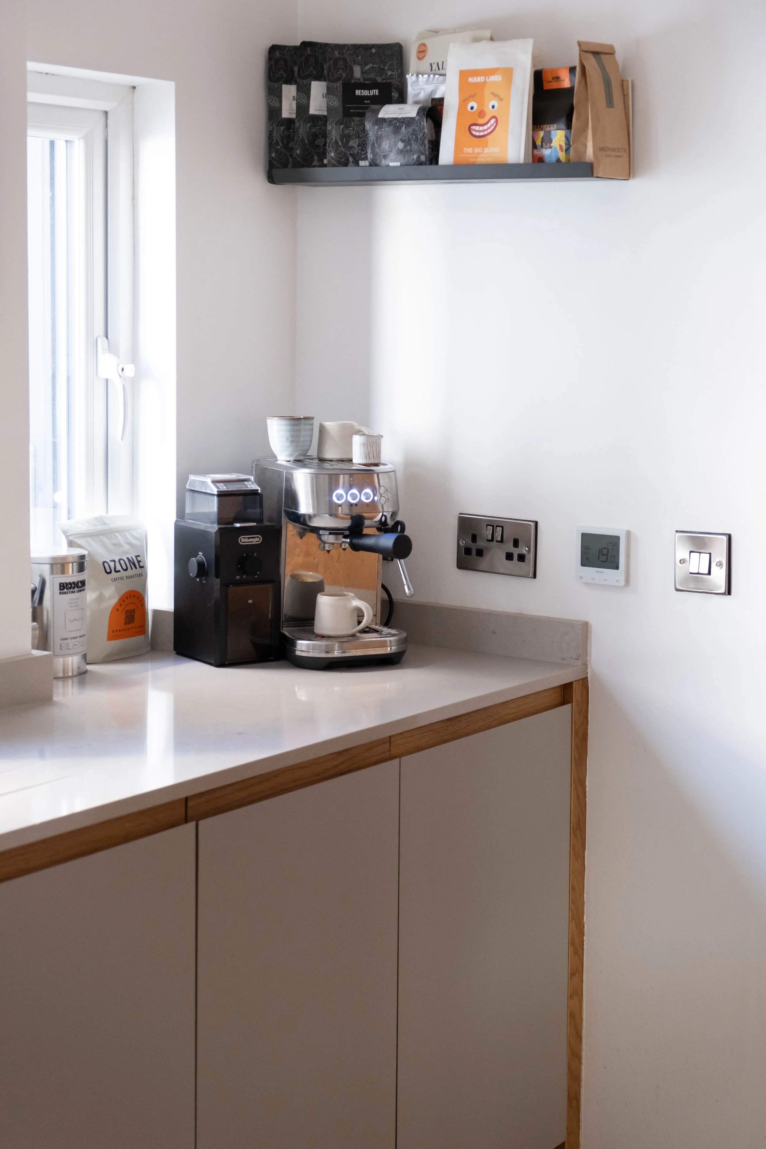 Countertop with a coffee machine, various cups, and coffee bags, with a white wall and electrical outlets in the background.