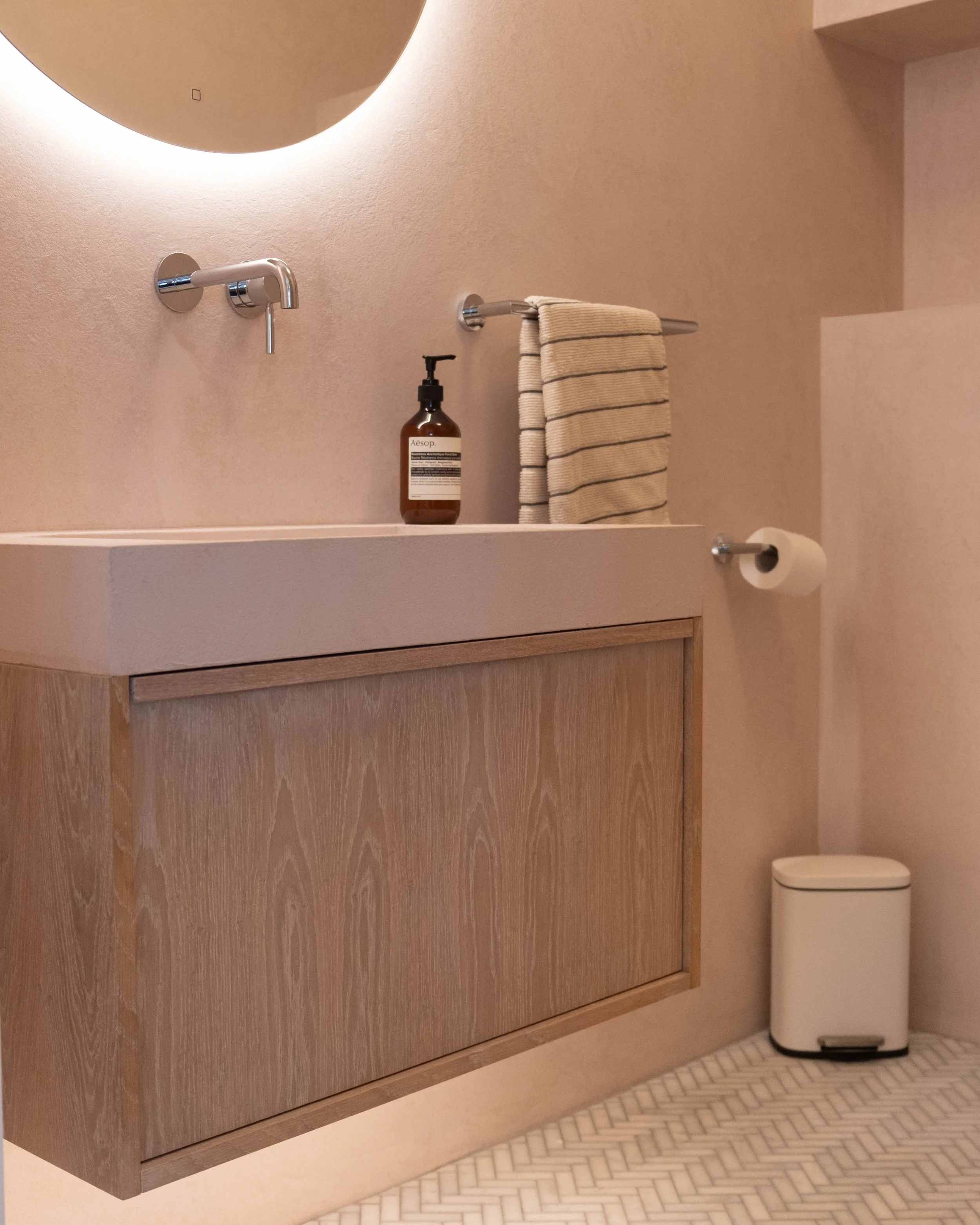A bathroom with a minimalist design including a wooden vanity with a beige countertop, a wall-mounted faucet, a bottle of Aesop hand soap, a striped beige and black towel on a silver towel bar, a rounded mirror with backlighting, a toilet paper holde