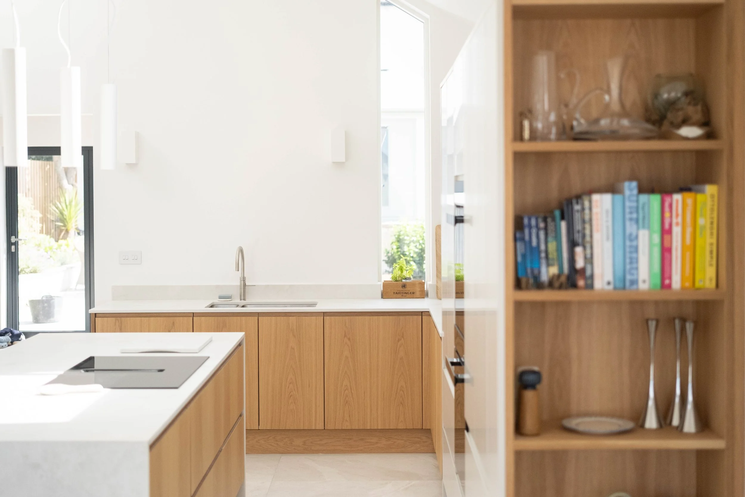 Modern kitchen with white walls, wooden cabinets, a white countertop, a black stovetop, a sink, a window, and a bookshelf with colorful books and decorative items.