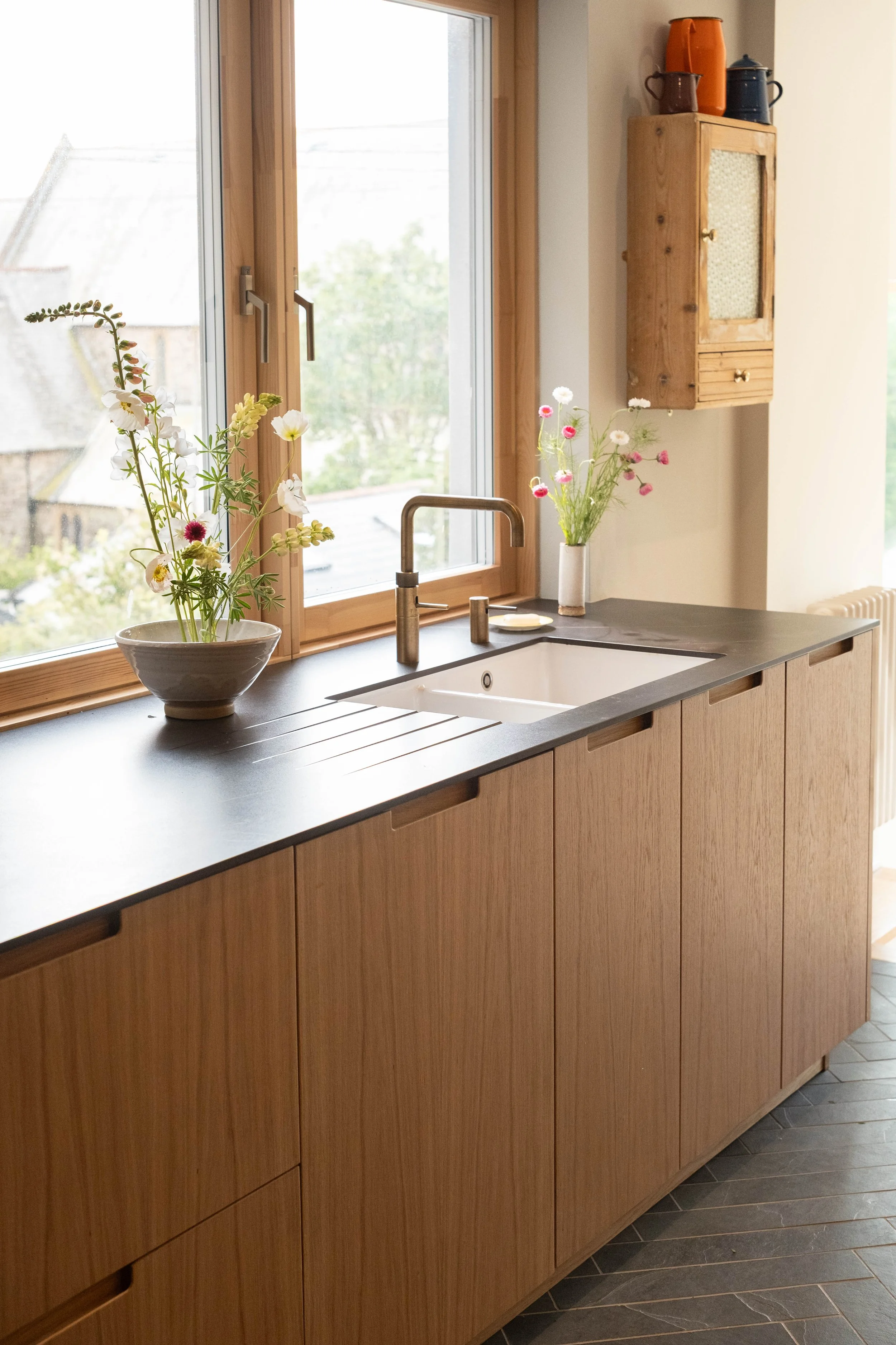 A kitchen with a black countertop, wooden cabinets, a stainless steel sink, a modern faucet, and two vases with flowers. A wooden cabinet mounted on the wall holds colorful pitchers and mugs. A window above the sink allows natural light in, with a vi