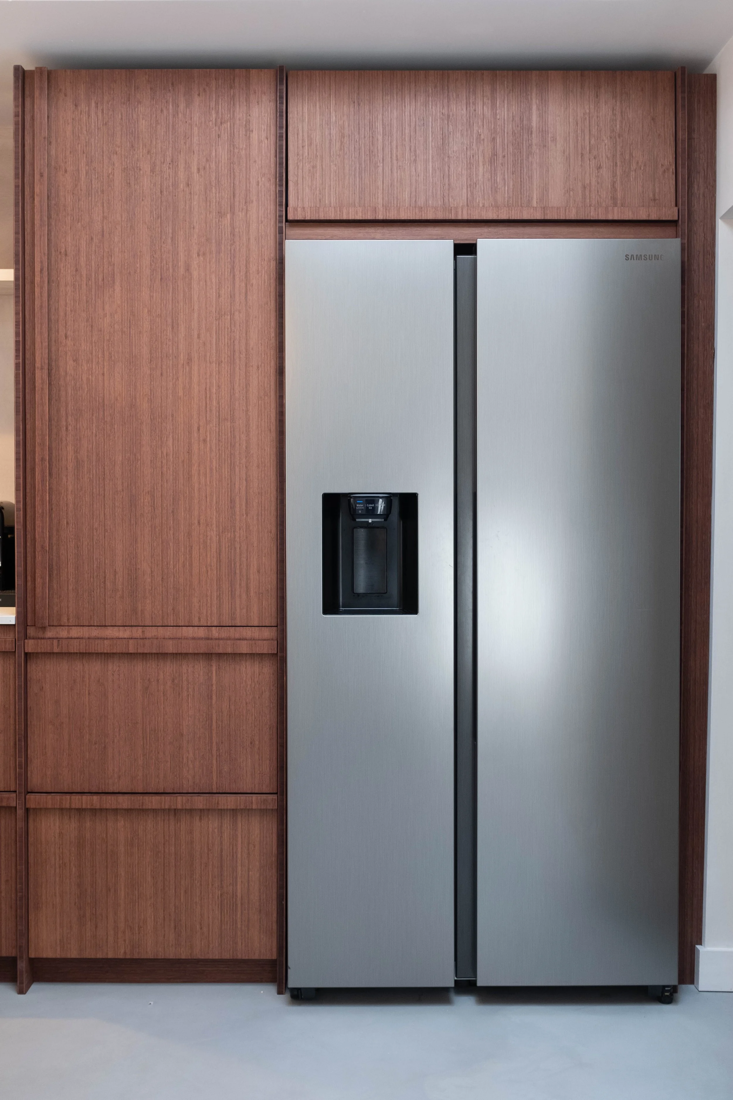 A stainless steel Samsung side-by-side refrigerator with a water and ice dispenser on the left door, placed between wooden cabinets in a kitchen.