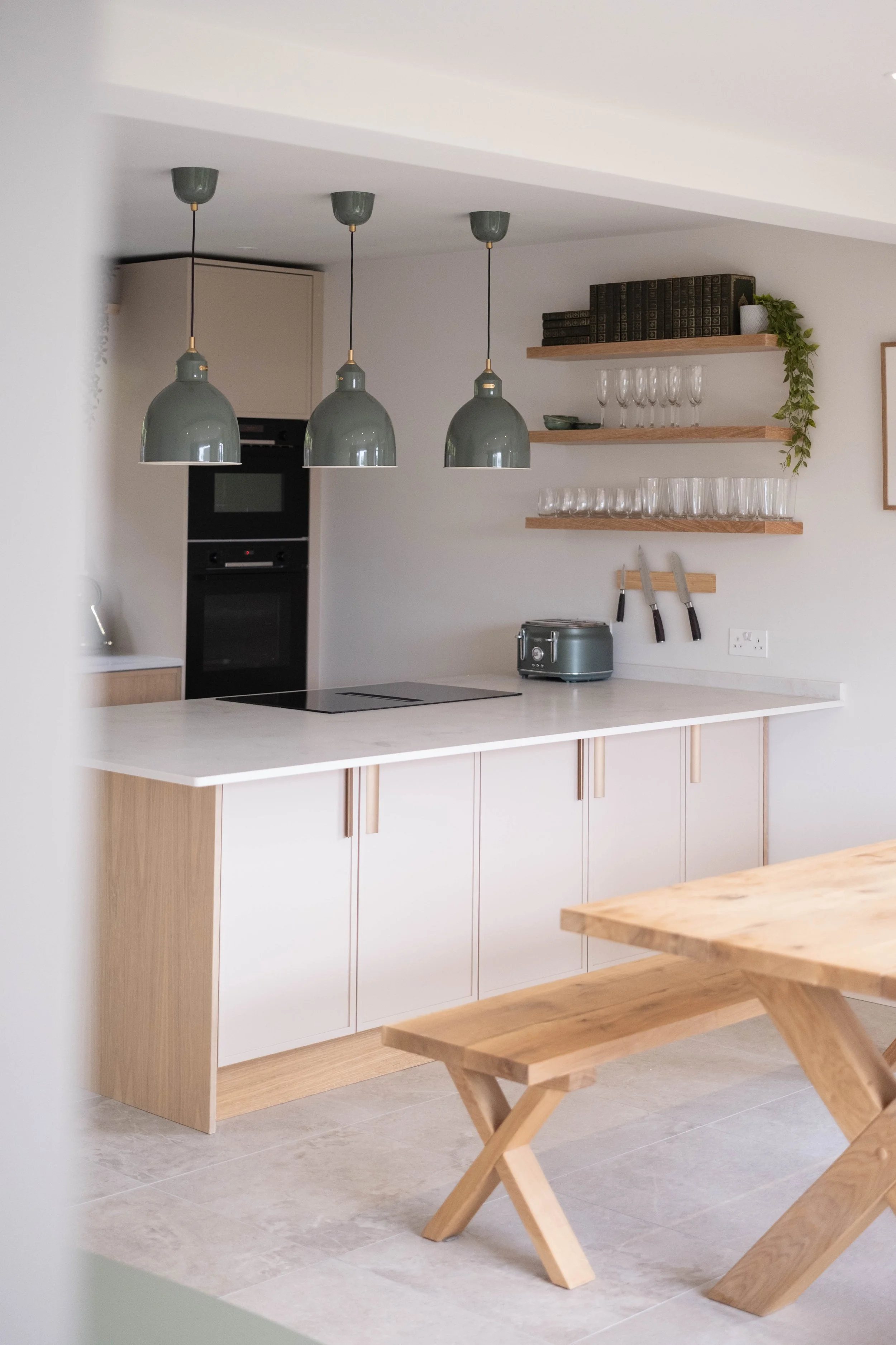 Modern kitchen with light wooden cabinets, white countertop, black stove, hanging green pendant lights, open wooden shelves with glasses, a toaster, and knives on a magnetic strip, and a wooden dining bench in the foreground.