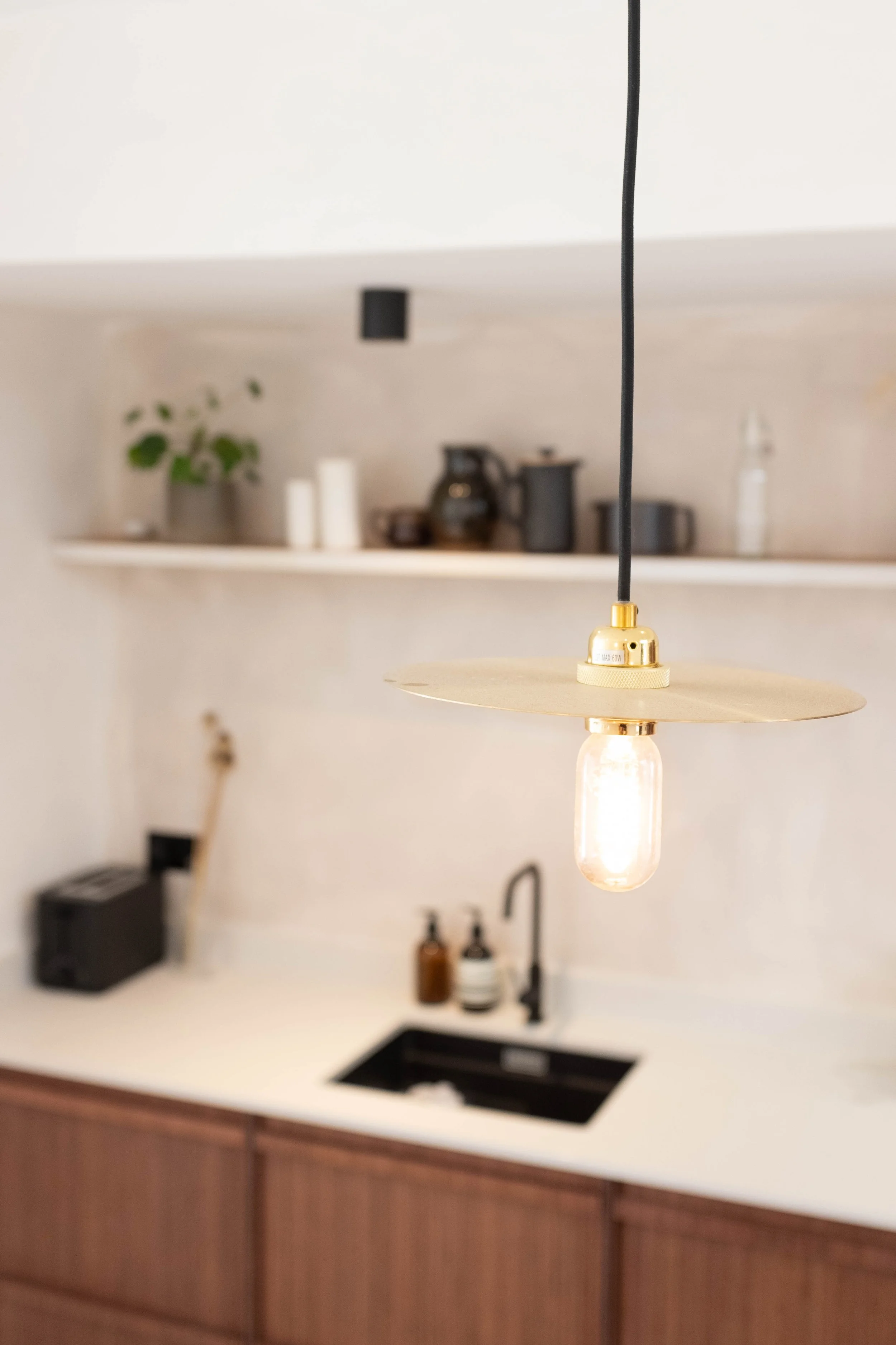 Close-up of a hanging pendant light with a black cord and gold fixture in a modern kitchen. In the background, a white shelf with decorative items and a small plant is visible. The kitchen counter has a built-in black sink, and a wooden cabinet with 