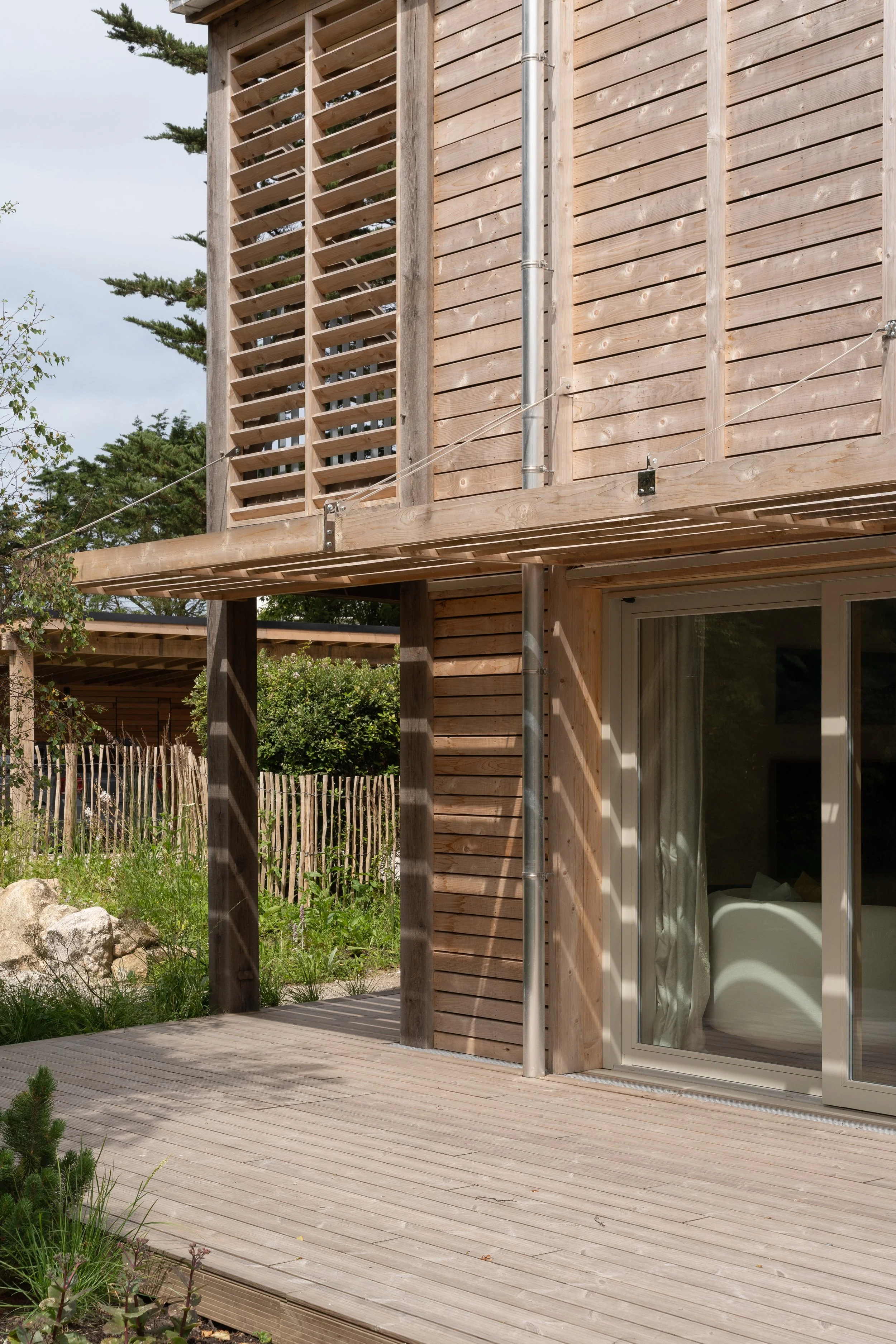 A wooden deck outside a house with a sliding glass door leading into a room. The house has vertical wooden siding and a metal drain pipe. A tree with pine branches can be seen in the background.