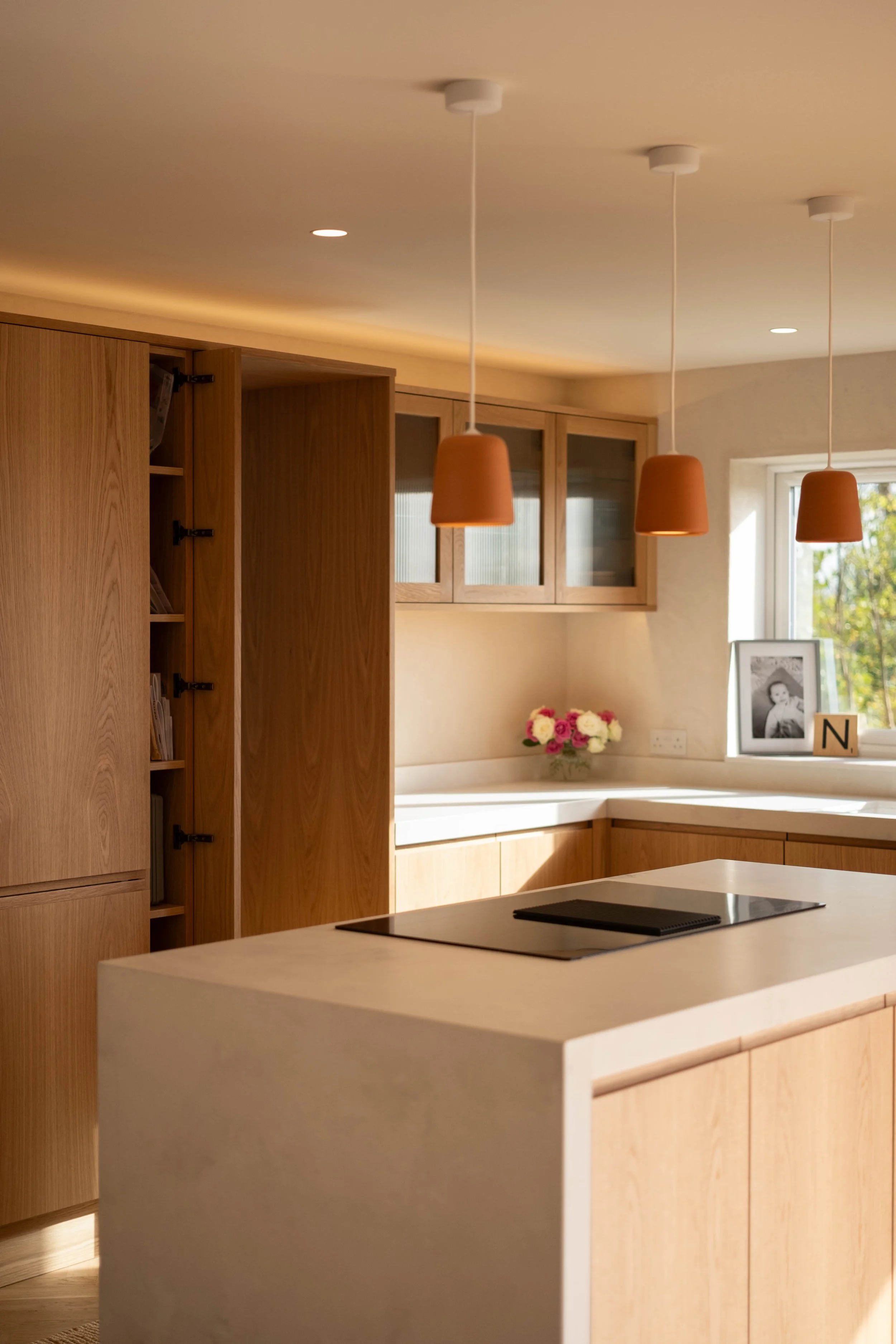 Modern kitchen with light wood cabinets and white countertops, featuring three terracotta pendant lights hanging above, a framed black-and-white photo on the windowsill, and a vase of pink and white flowers.
