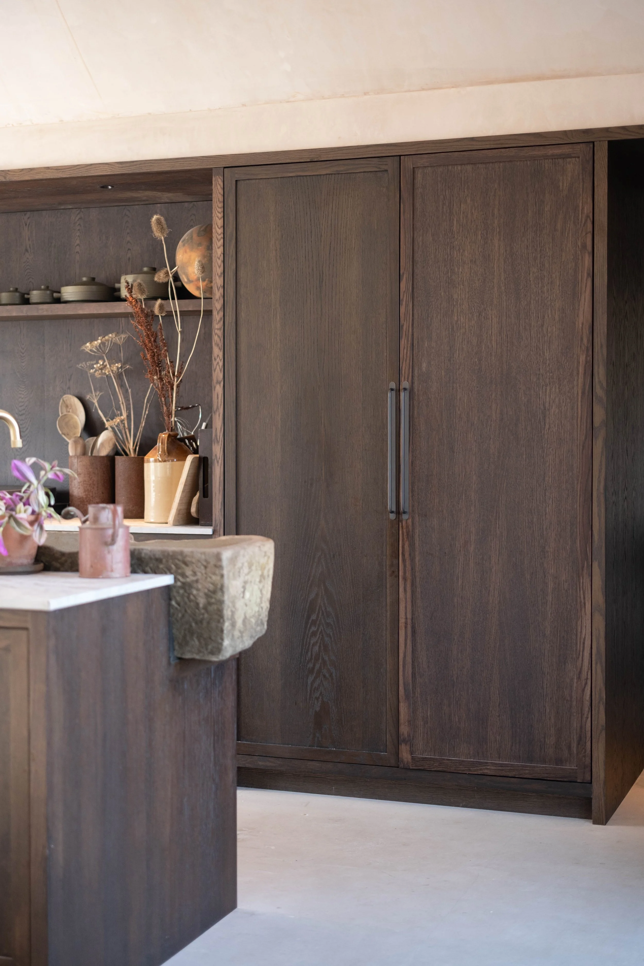 A kitchen with wooden cabinets and a table with vases, dried flowers, and pottery. There are open shelves with dishes and a globe.