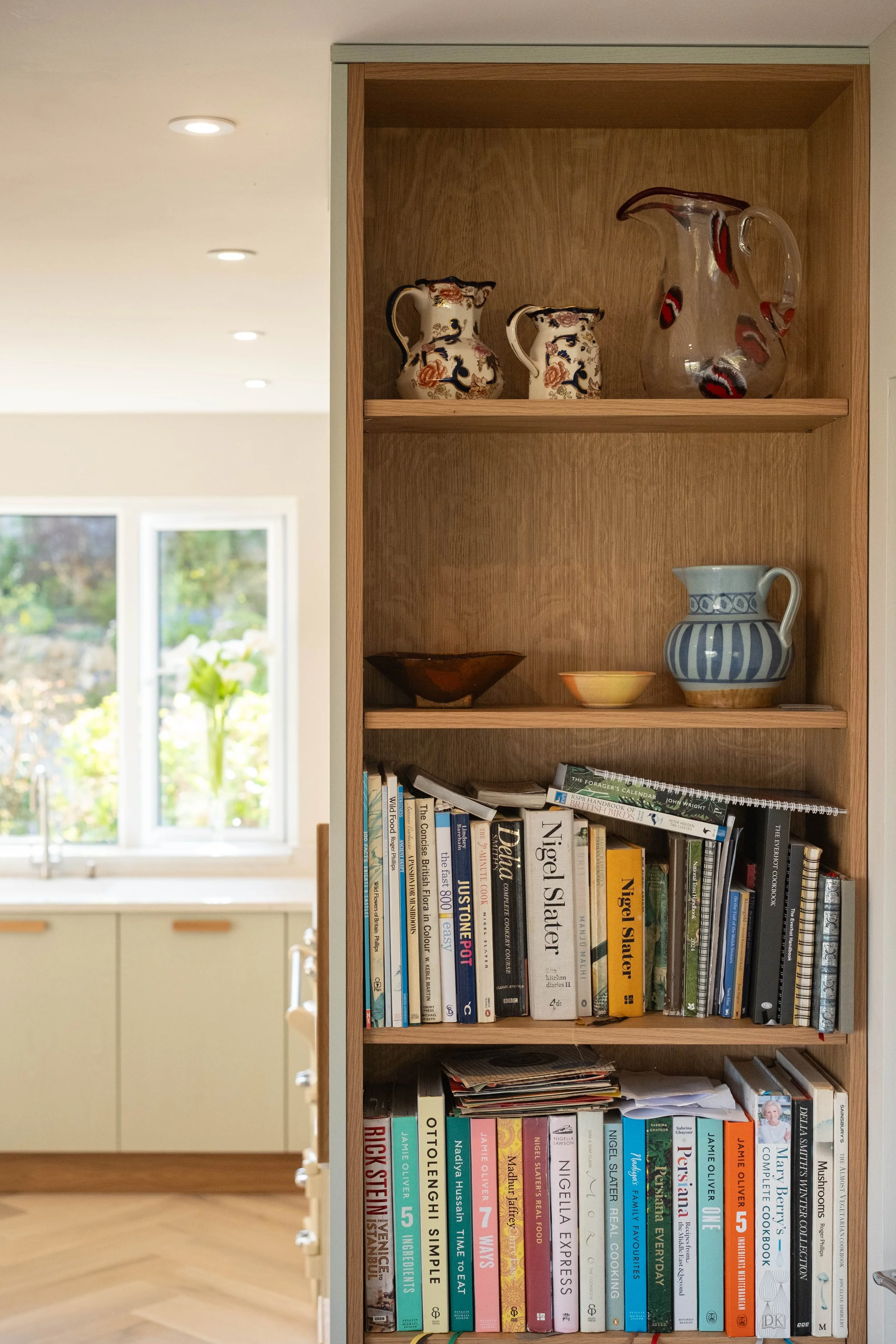 A wooden bookshelf with decorative vases on top, a bowl and a pitcher, and a collection of cookbooks and magazines on the lower shelves, with a kitchen and a window showing outdoor greenery in the background.