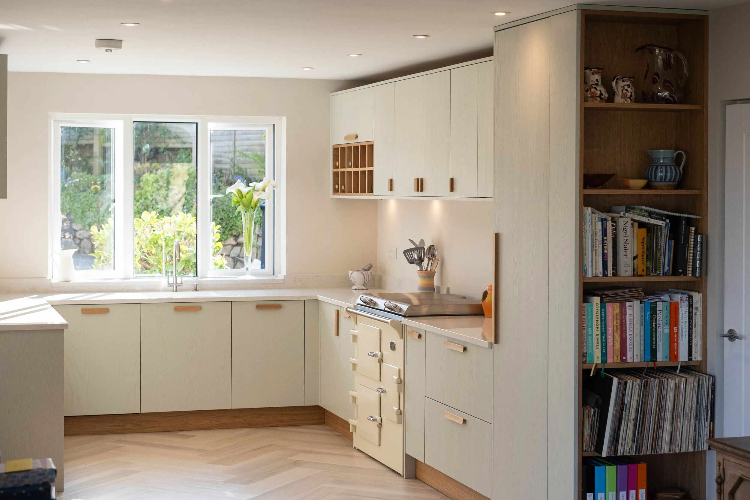 A modern kitchen with white cabinets, a window with a view of a garden, and a bookshelf filled with books.