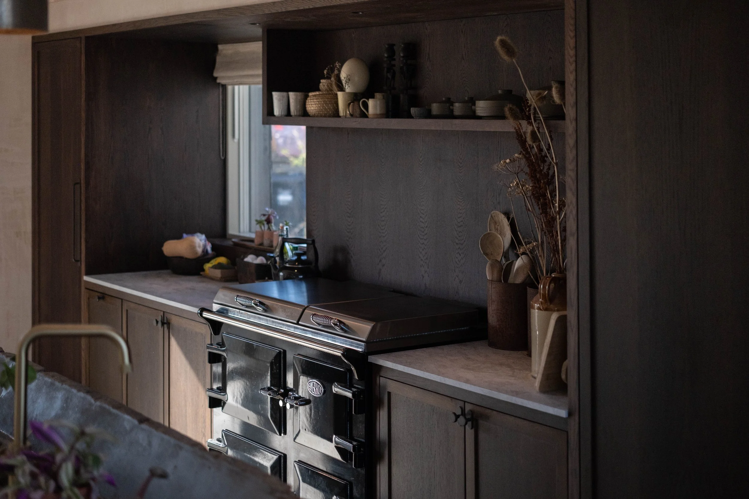 A kitchen with dark wooden cabinets and a black stove, with a window in the background, decorative dishes and plants on the shelves, and dried flowers in a container on the counter.