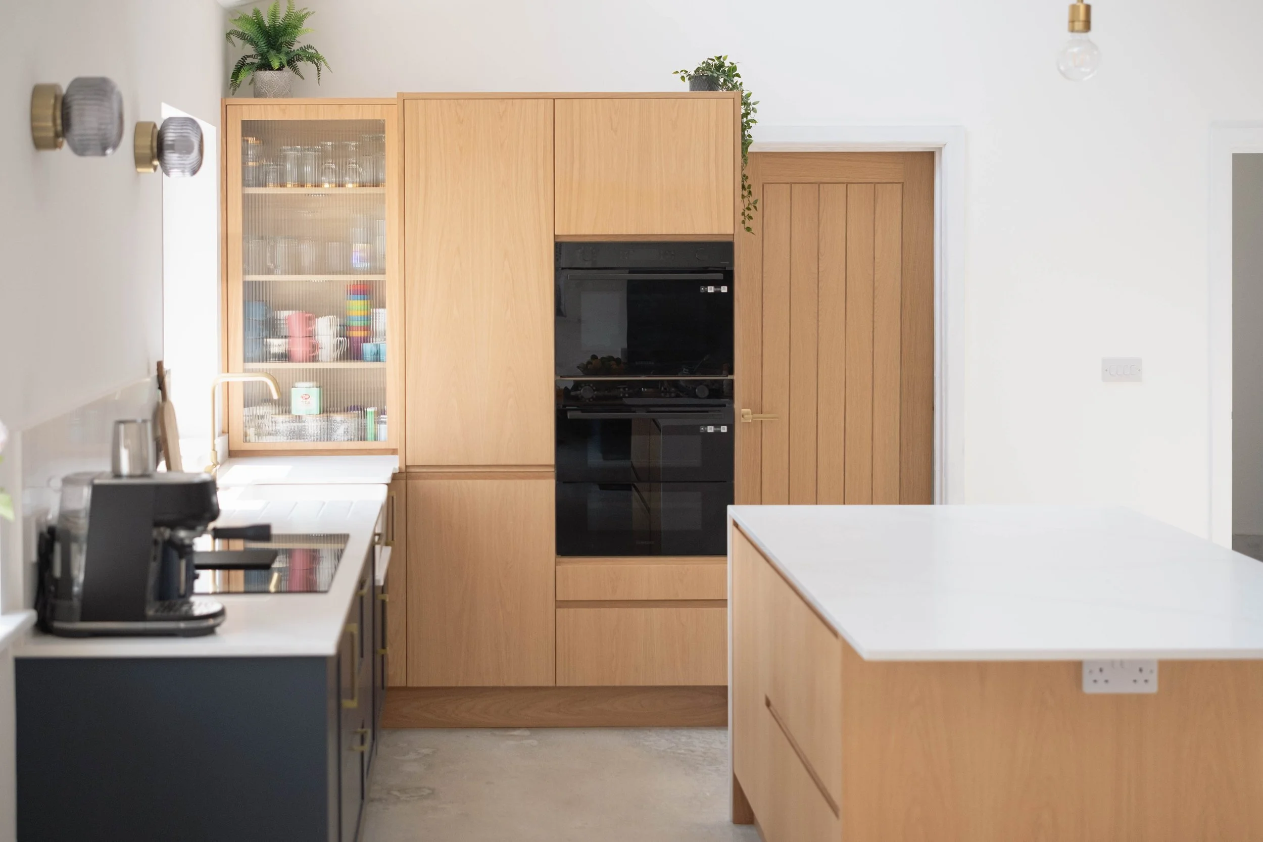 Modern kitchen featuring light wood cabinetry, black built-in oven, white countertops, and decorative plants on top of cabinets.
