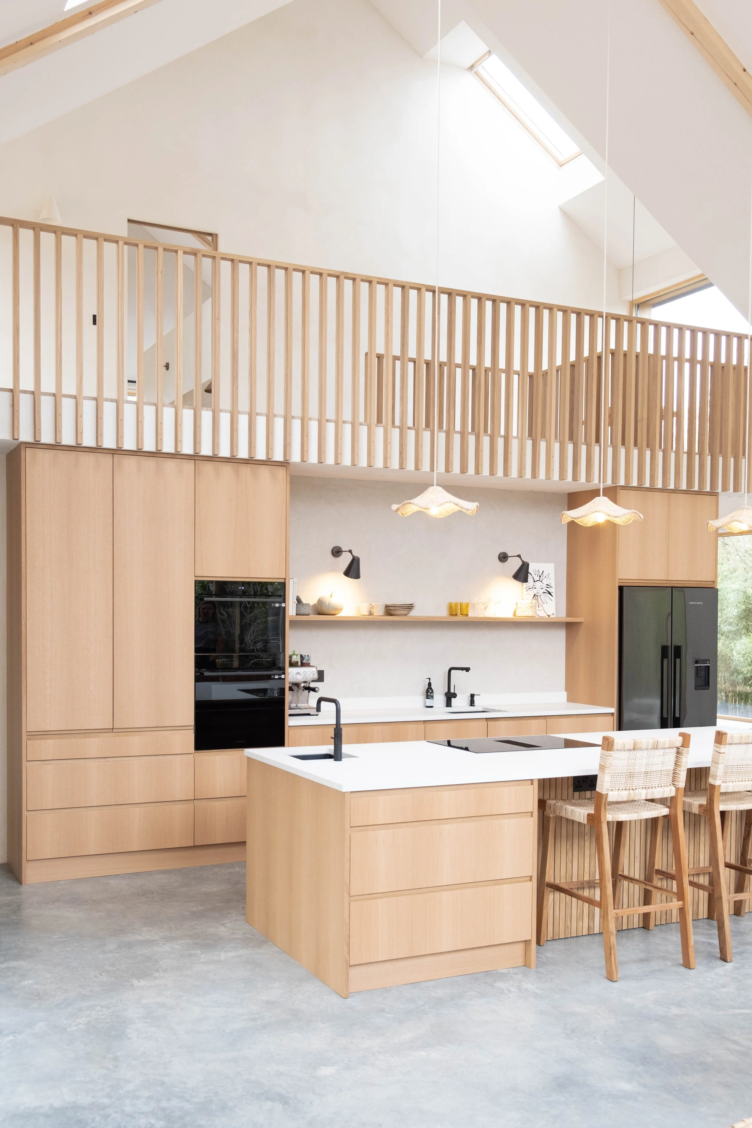 Modern kitchen with light wood cabinets, white countertops, black appliances, and two wicker chairs at the counter. The upper area has a wooden railing and skylight windows.