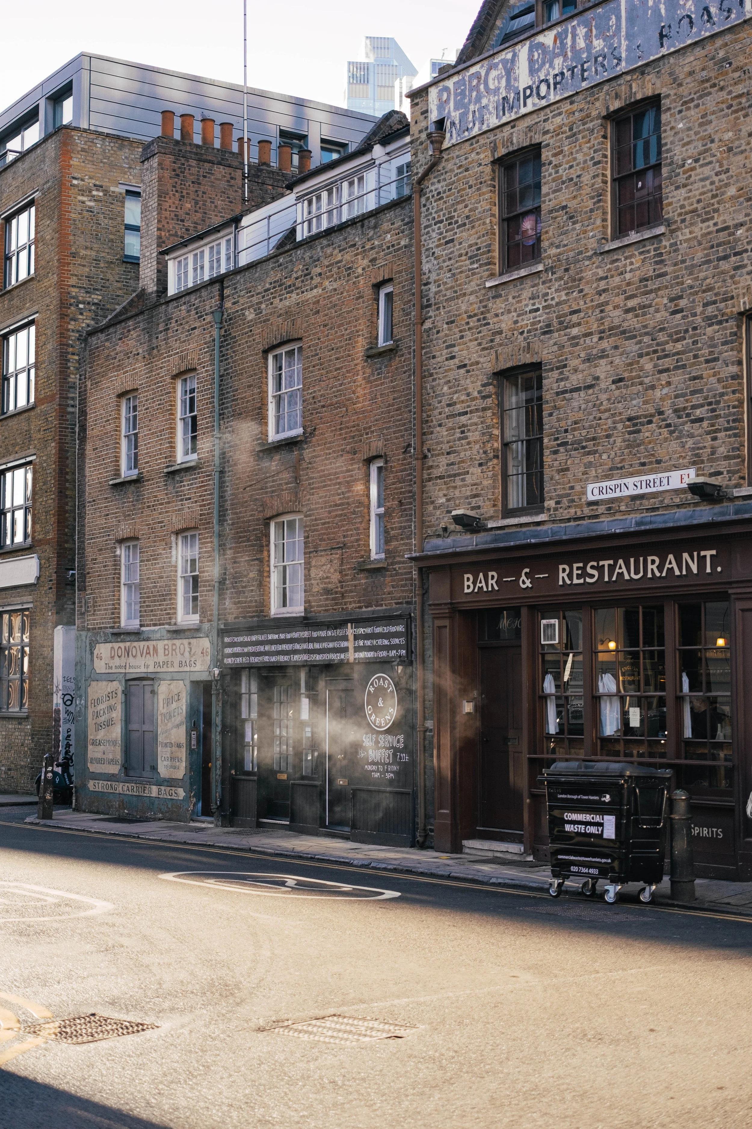 Street view of brick buildings, including a bar and restaurant with large glass windows, a street sign reading 'Crispin Street', a waste management bin, and a sidewalk with a crosswalk.