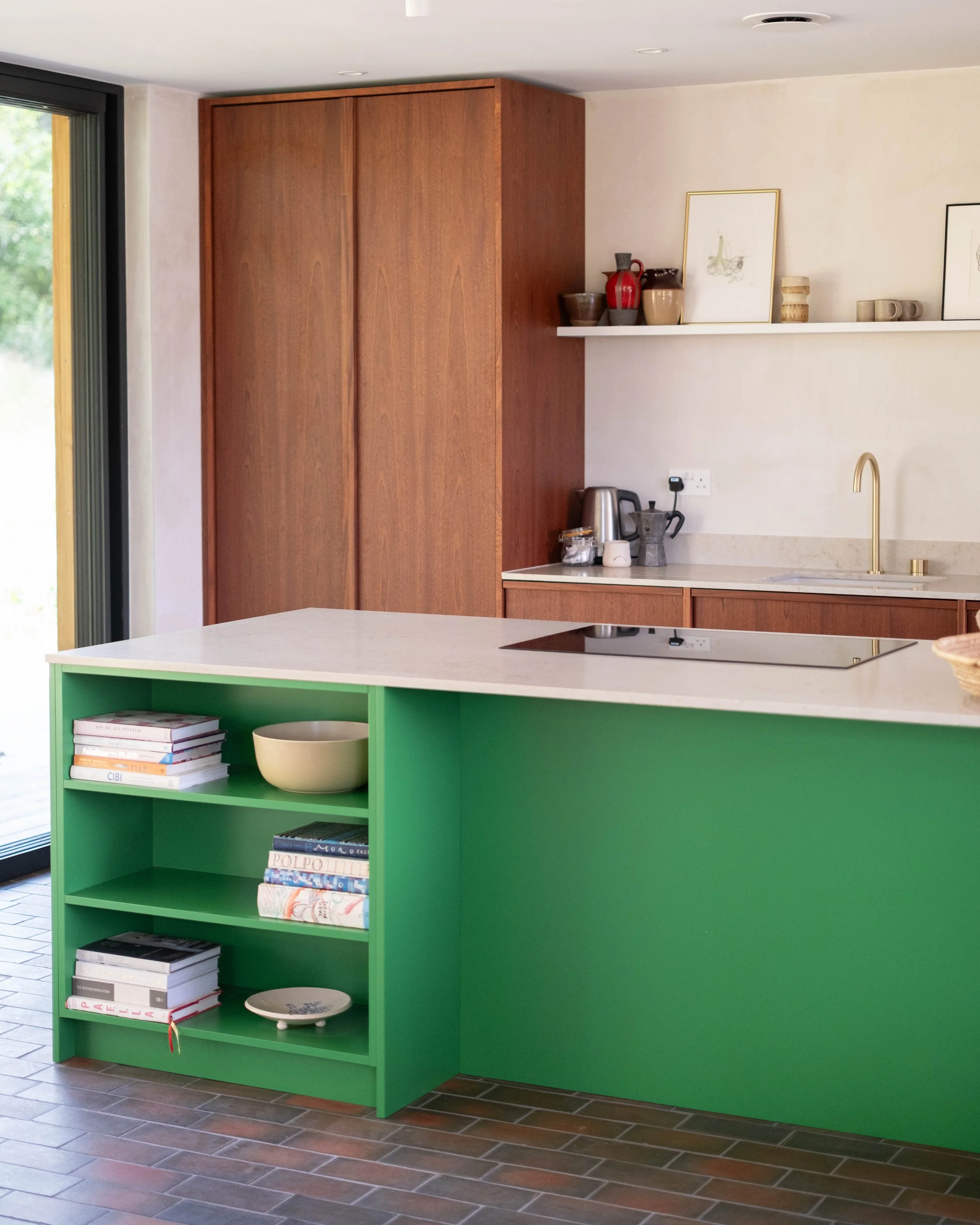 A kitchen with a green island, wooden cabinets, a floating shelf with decorative items, and a sliding glass door leading outside.